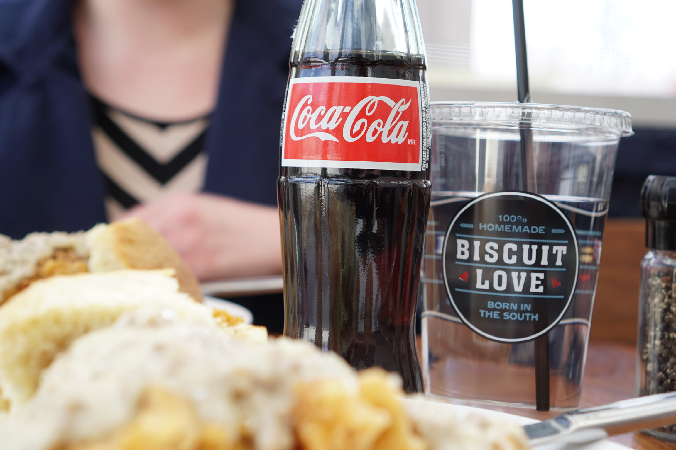 A bottle of Coca-Cola and a clear plastic cup labeled "Biscuit Love" on a table with plates of food, and a person sitting in the background.