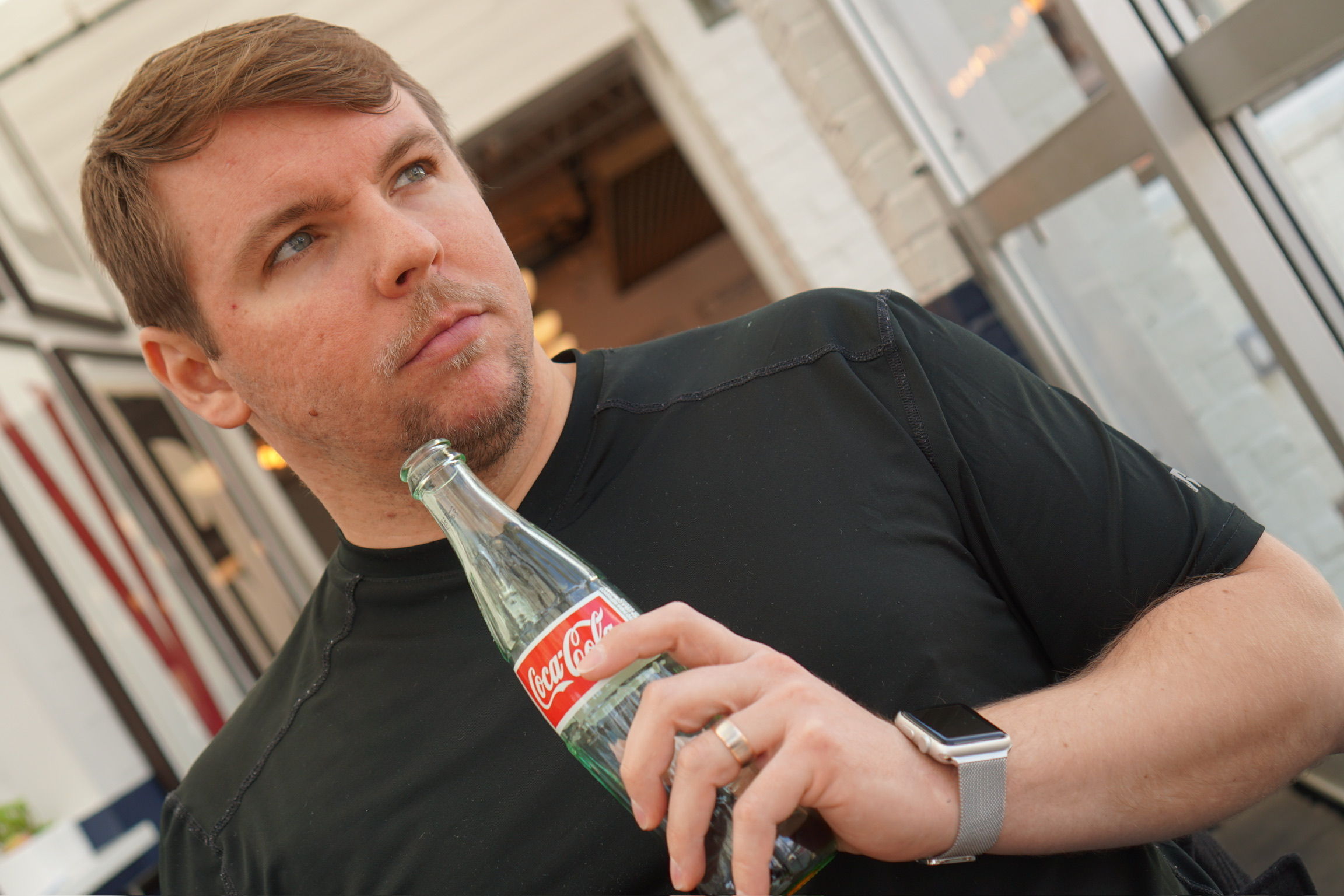A man in a black shirt holding a Coca-Cola glass bottle and wearing a smartwatch looks to the side inside a bright room.