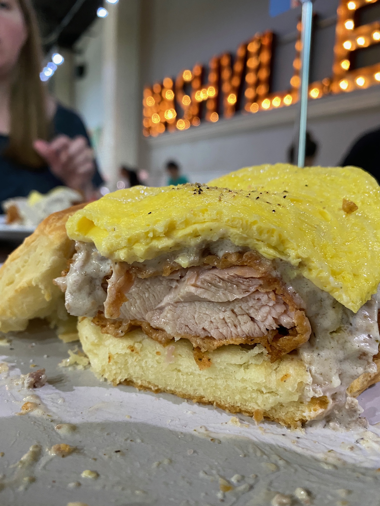 A close-up of a biscuit sandwich with fried chicken, yellow egg, and gravy, with a blurred restaurant sign reading "NASHVILLE" in the background.