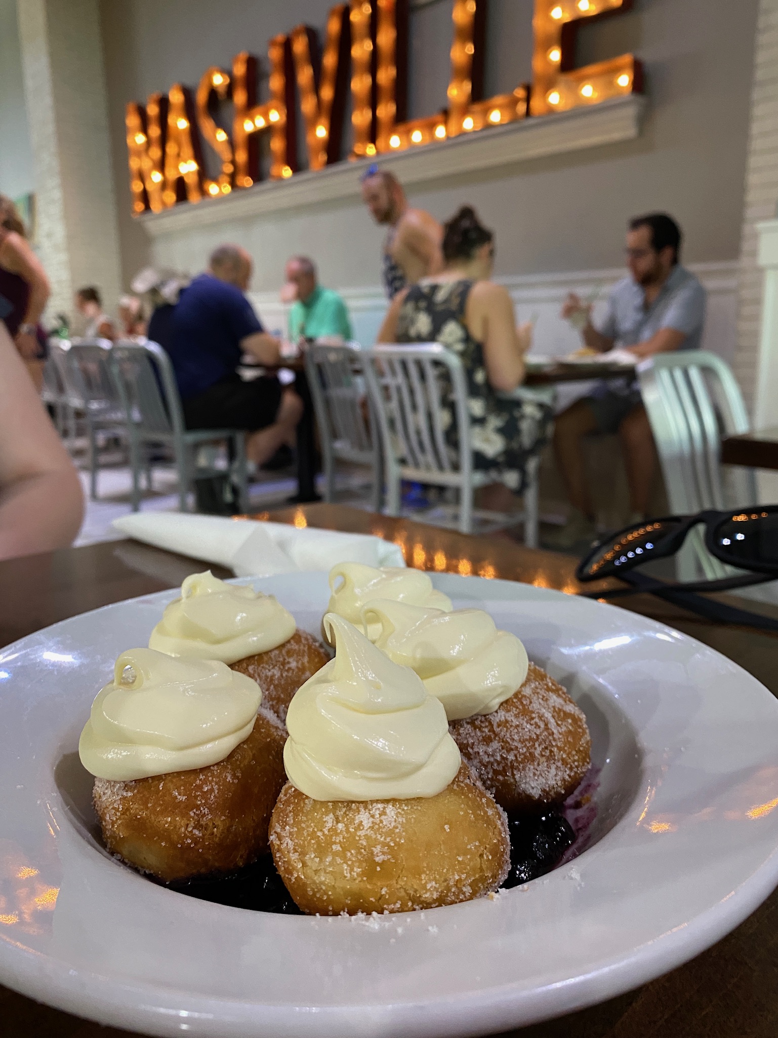 A plate of sugared donuts with cream topping sits on a table in a restaurant, with people dining and a lit "NASHVILLE" sign in the background.