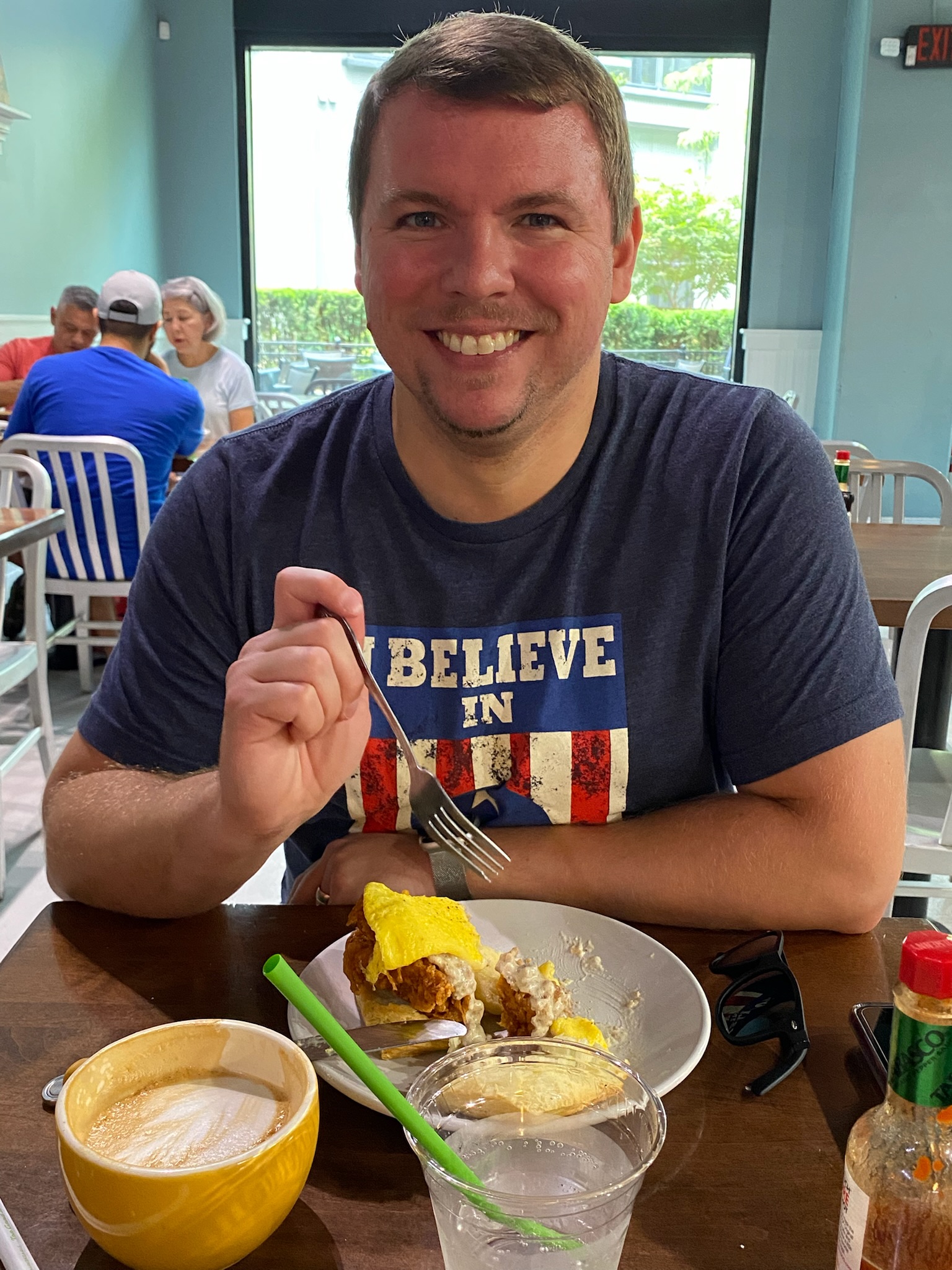 A man sits at a table in a restaurant, smiling at the camera while holding a fork over a plate of food, with a cup of coffee, a glass of water, and hot sauce bottles on the table.