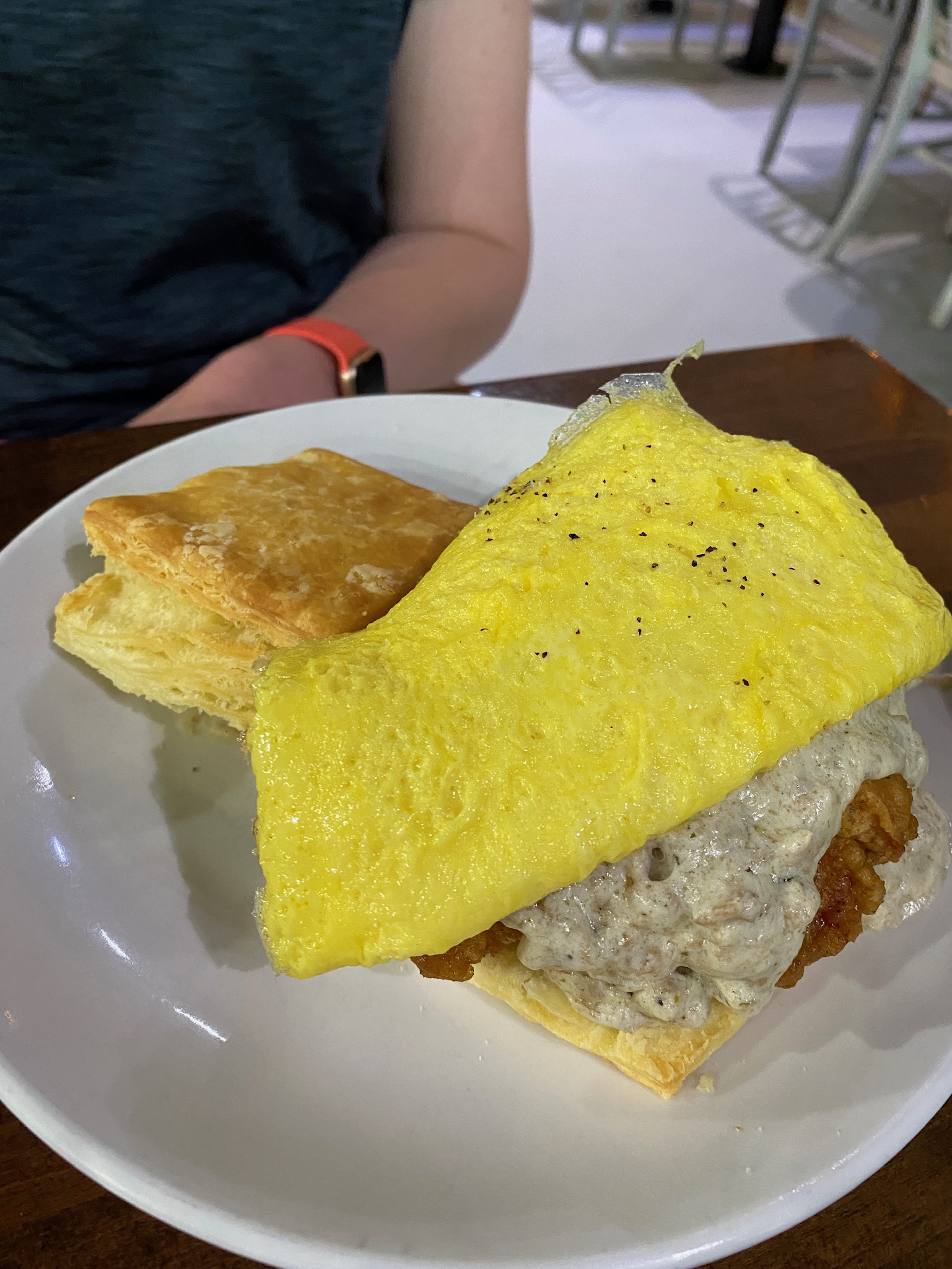 A biscuit sandwich with fried chicken, sausage gravy, and an omelette on top is served on a white plate; a person sits in the background.