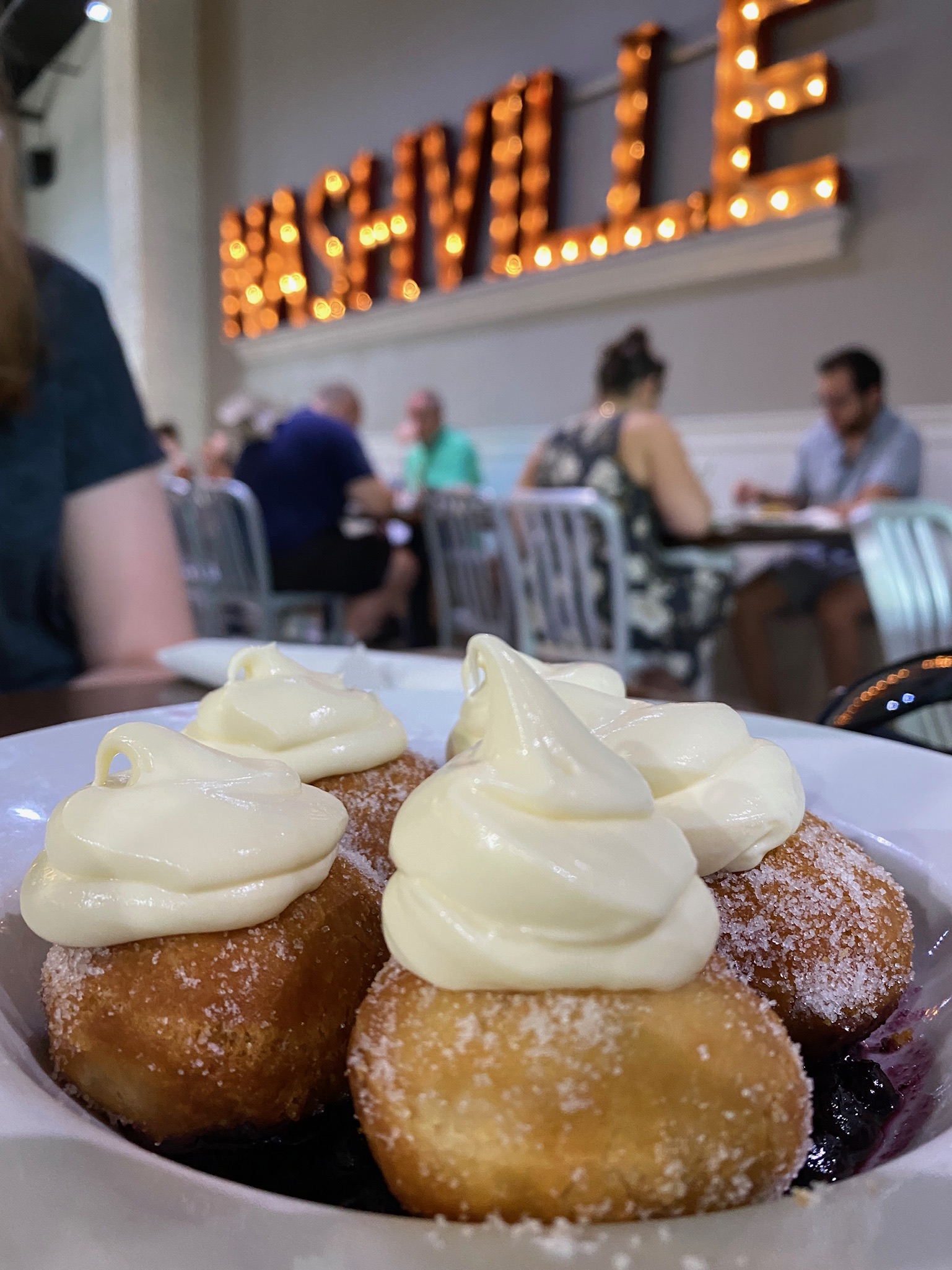 Four sugar-dusted doughnuts topped with cream are served in a bowl with a blurred restaurant background and a large illuminated "NASHVILLE" sign on the wall.