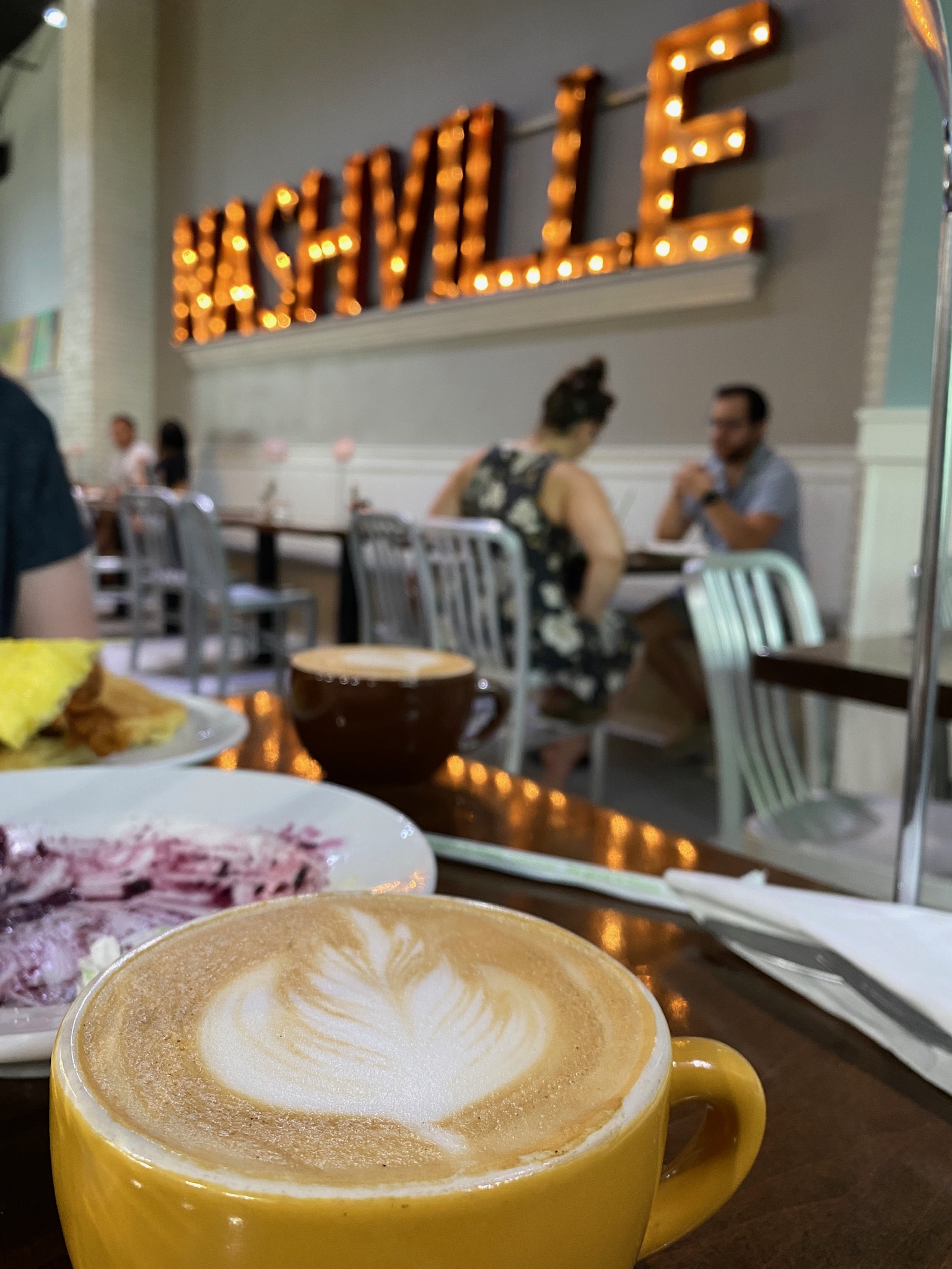 A latte in a yellow cup on a table with food, with people dining in the background and a lit “NASHVILLE” sign on the wall.