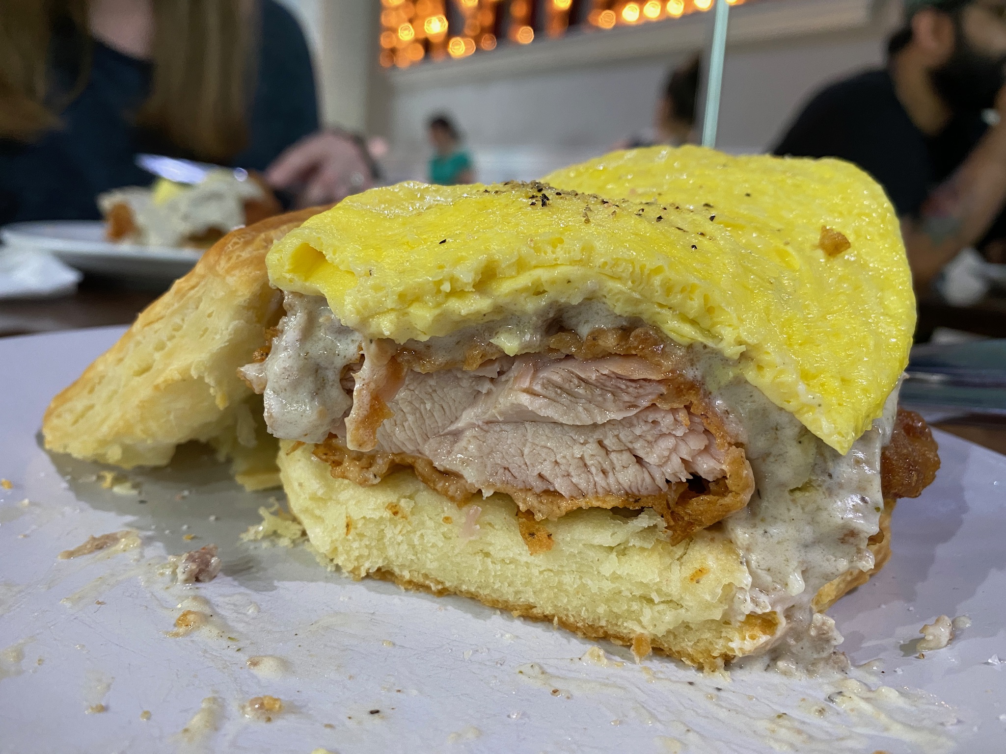 A biscuit sandwich filled with fried chicken, egg, and sausage gravy sits on a plate in a restaurant setting.