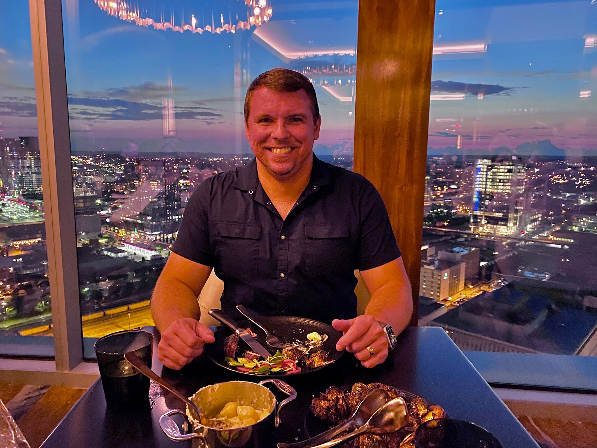 A man sits at a restaurant table with food, smiling at the camera. Large windows behind him show a cityscape at sunset with lights and tall buildings.