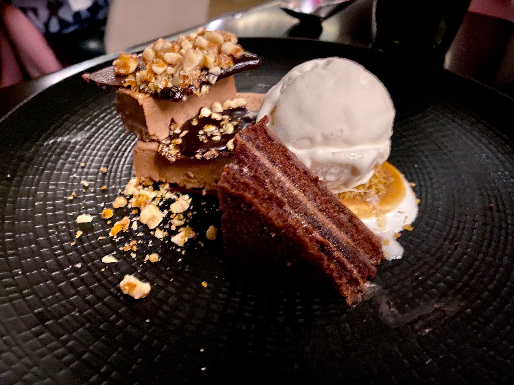 A dessert plate with a scoop of vanilla ice cream, a slice of chocolate cake, and a layered chocolate and nut confection, garnished with crushed nuts.
