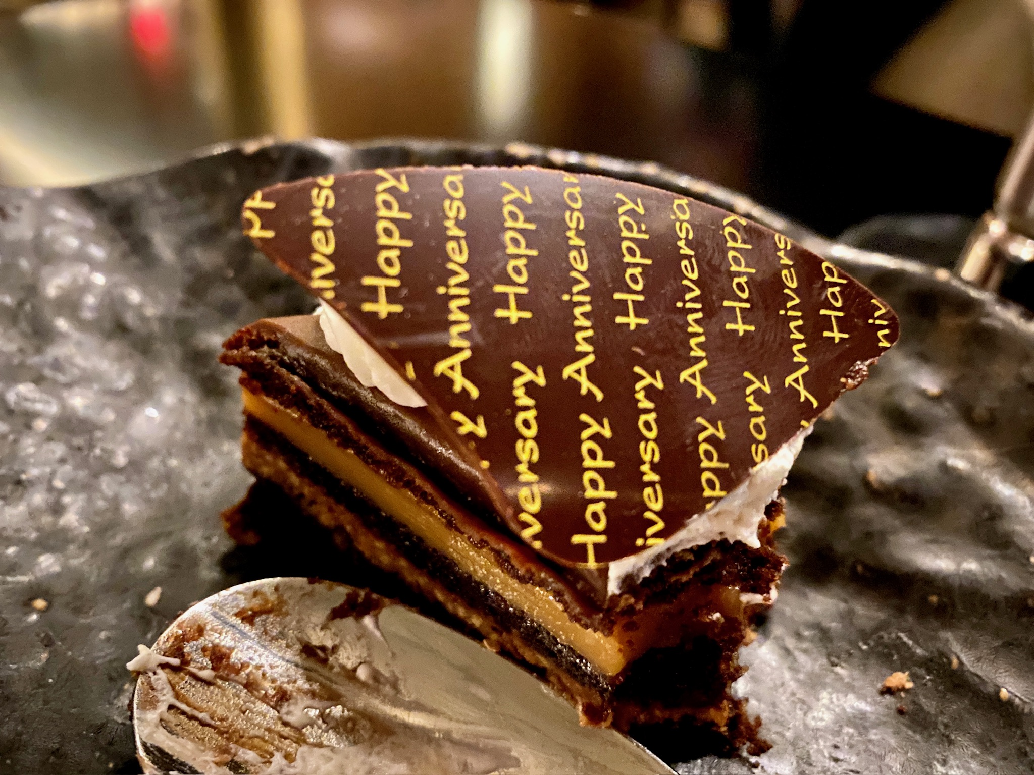 A partially eaten layered chocolate cake with a decorative chocolate topper that reads "Happy Anniversary" on a dark plate.