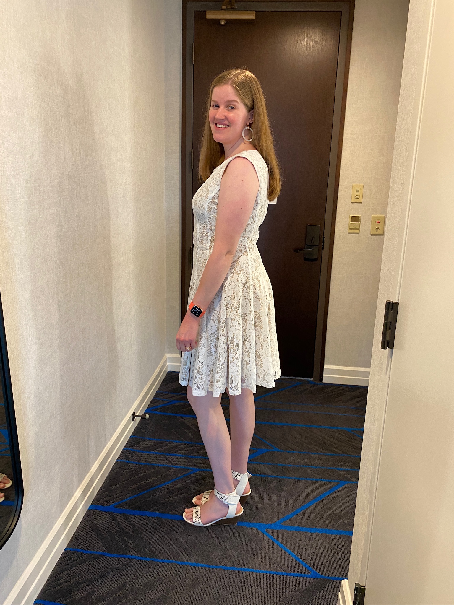 A woman in a white lace dress and white heels stands sideways in a hallway, smiling at the camera. She wears hoop earrings and a smartwatch.