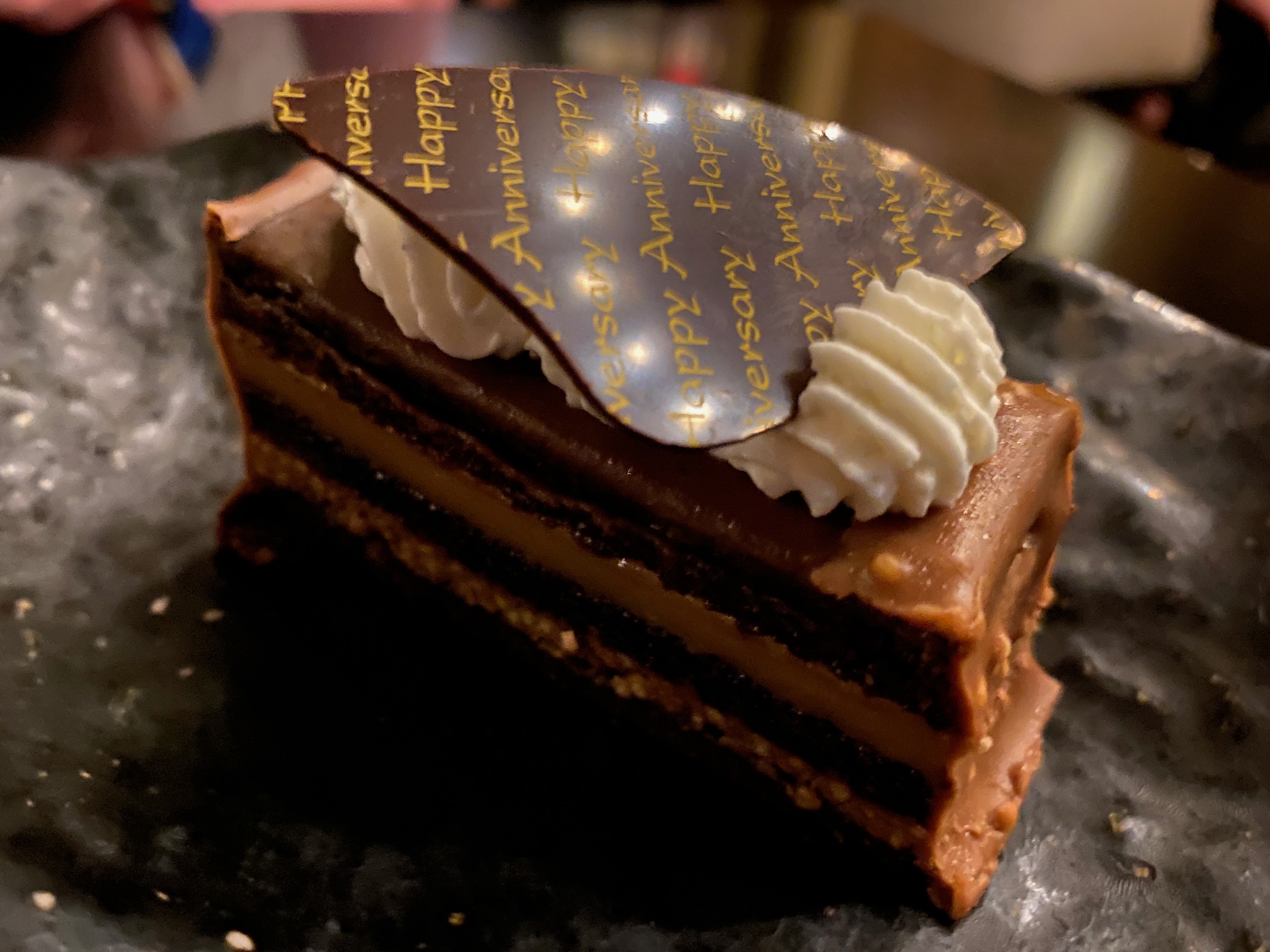 A slice of layered chocolate cake with cream and a decorative chocolate piece reading "Happy Anniversary" on a dark plate.