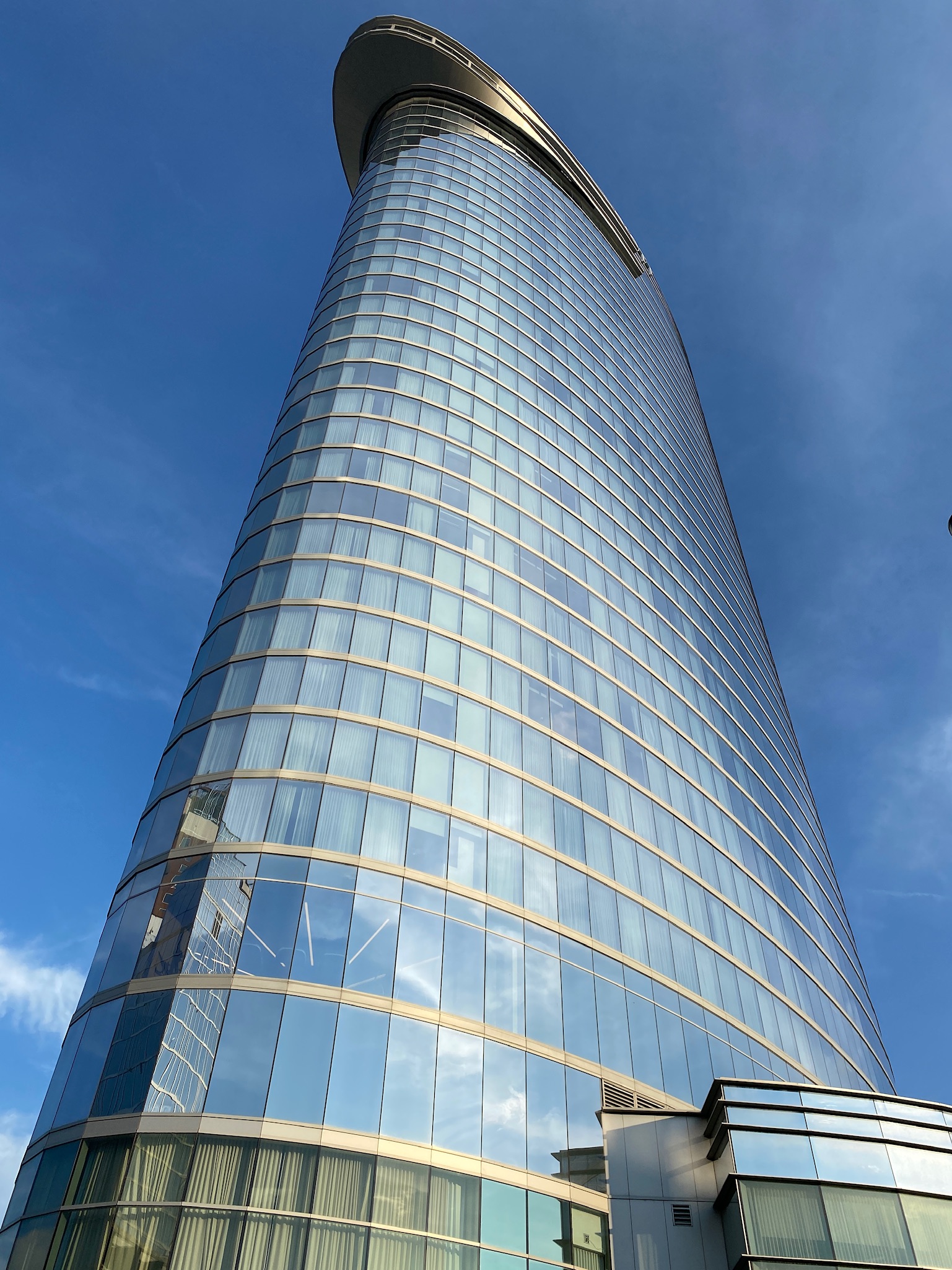 Tall modern glass skyscraper with a curved top against a blue sky, viewed from a low angle.