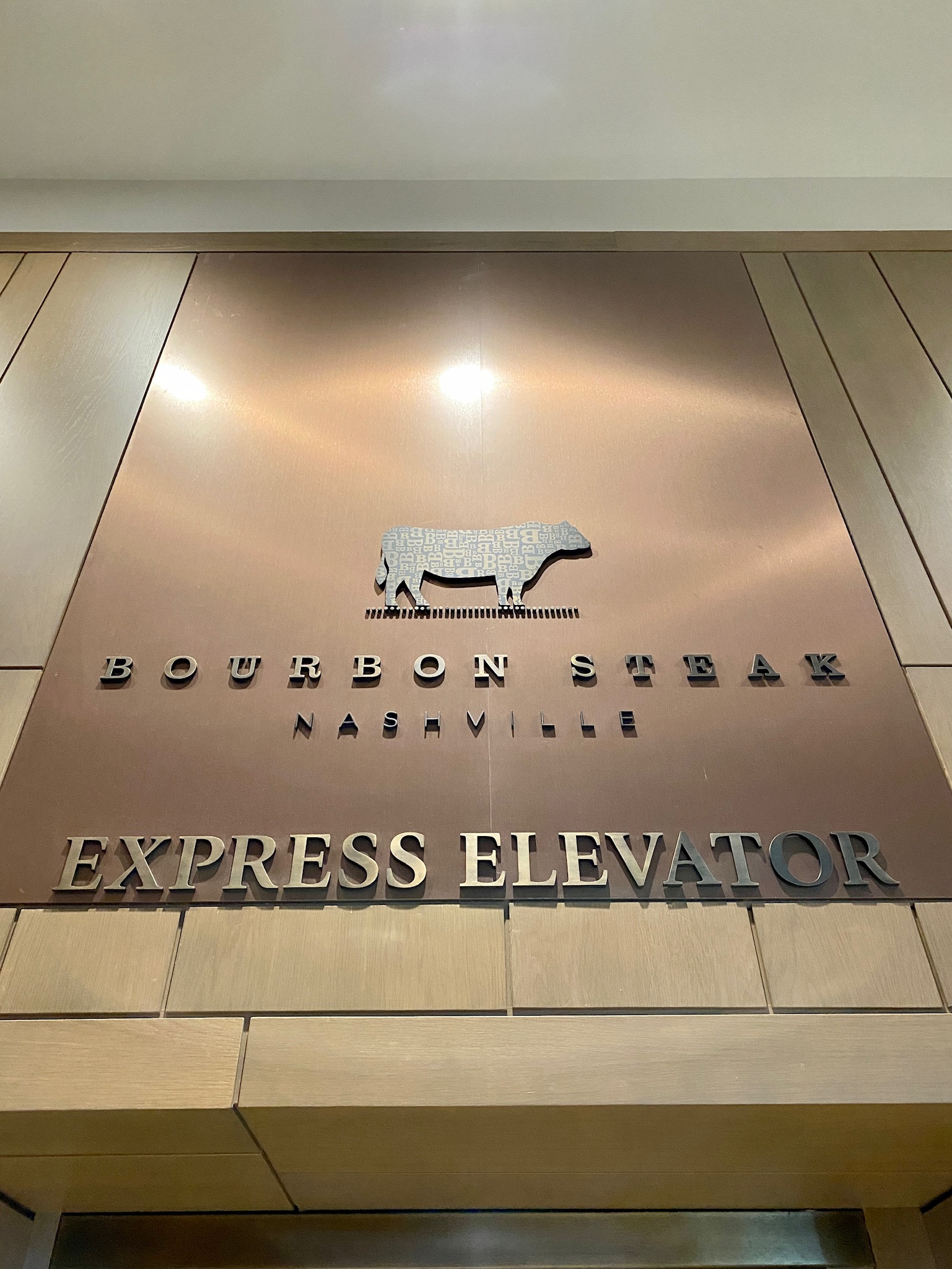 Sign for Bourbon Steak Nashville above an express elevator, featuring a stylized cow graphic and metallic lettering on a bronze background.