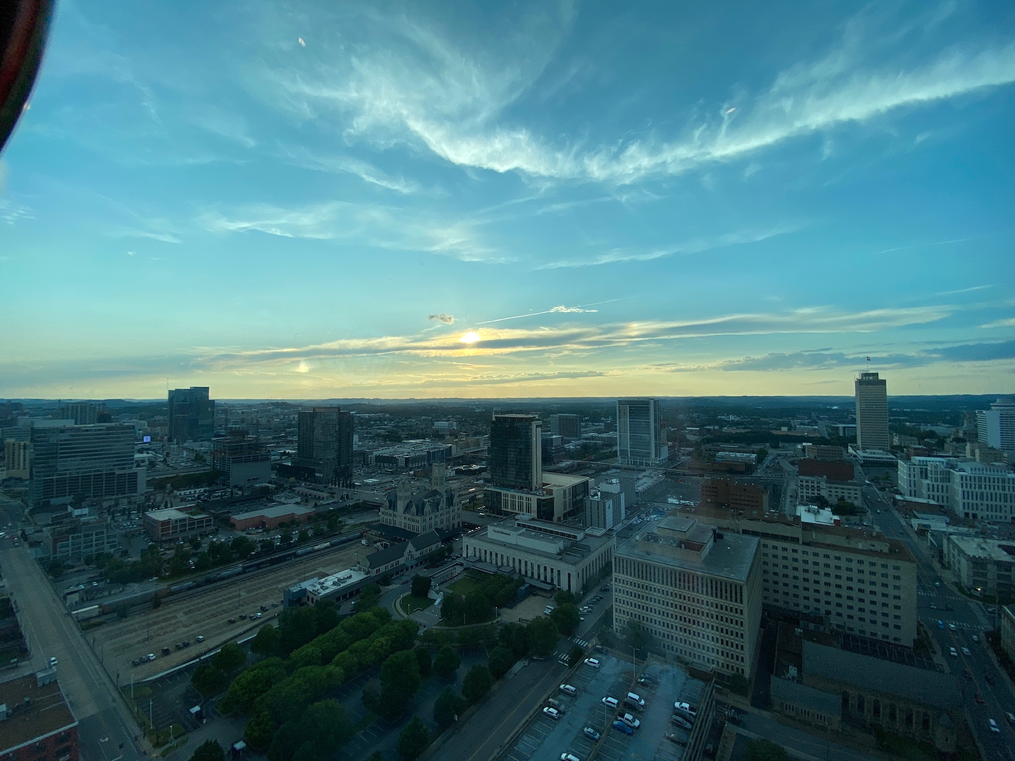 A cityscape view at sunset with buildings, parking lots, and tree-lined streets under a partly cloudy sky.