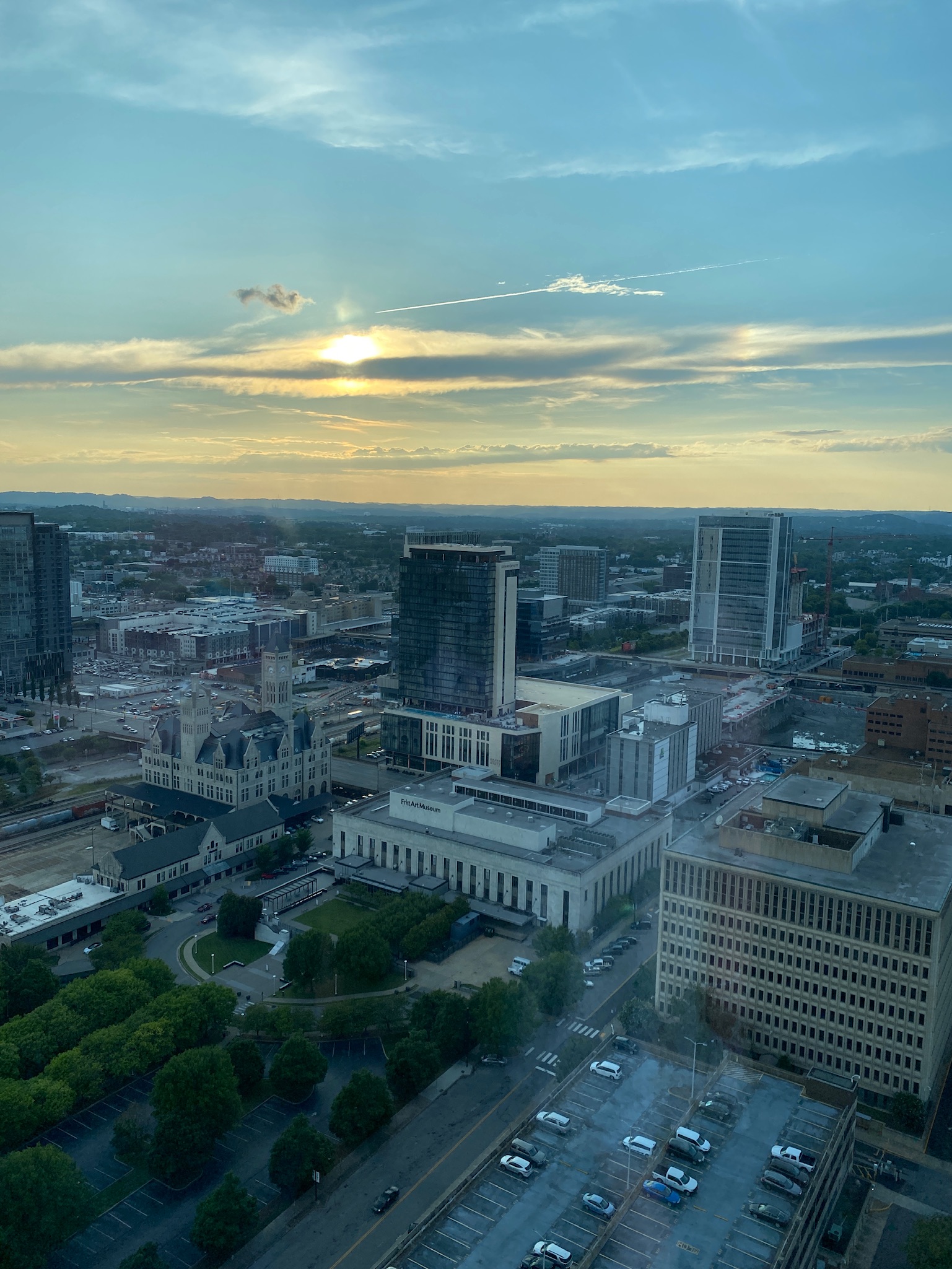 A cityscape at sunset with tall buildings, a parking lot, and cloudy sky; sunlight is visible near the horizon.