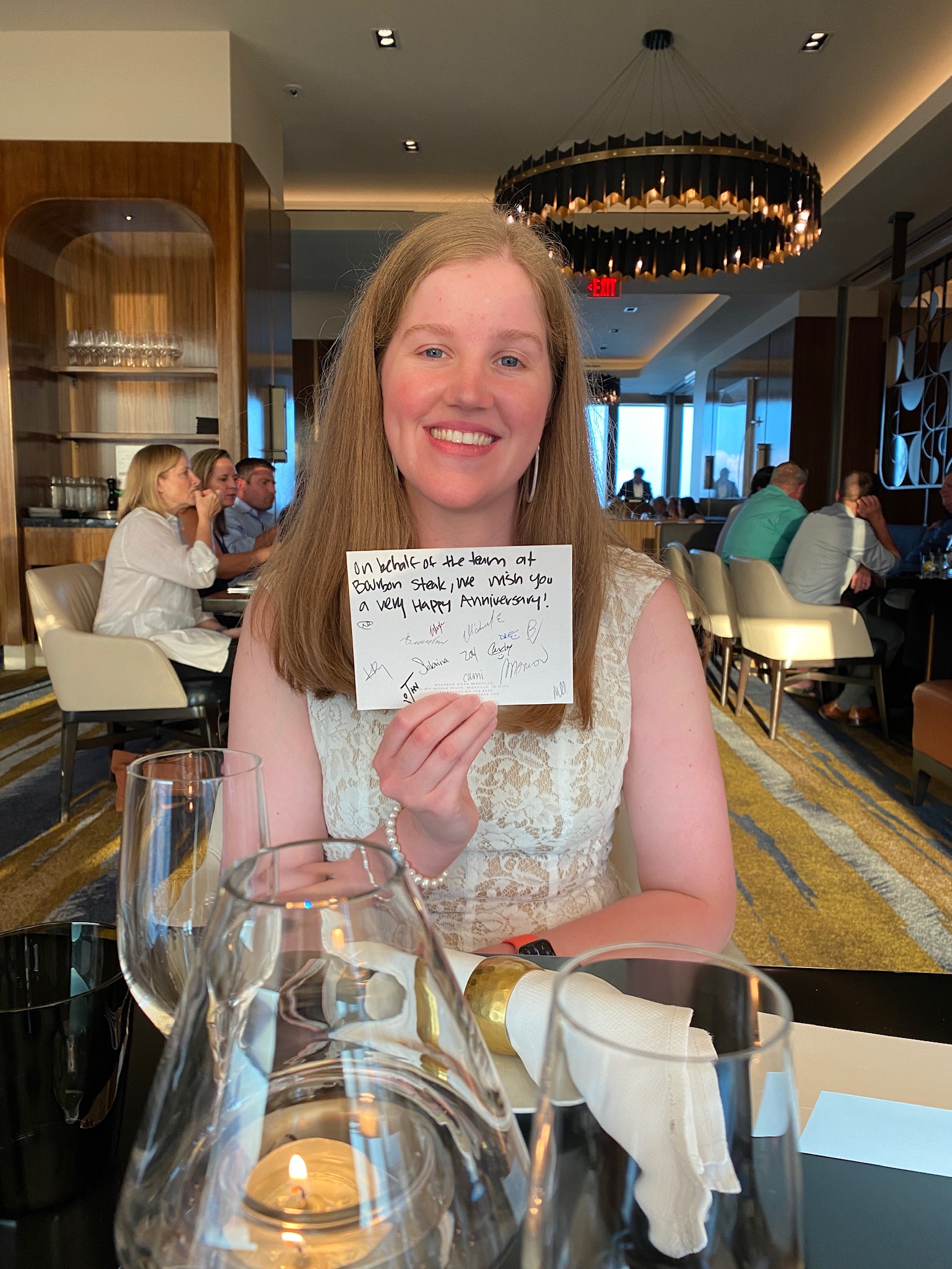 A woman in a white dress sits at a restaurant table, holding up a handwritten note and smiling at the camera. Other diners are visible in the background.