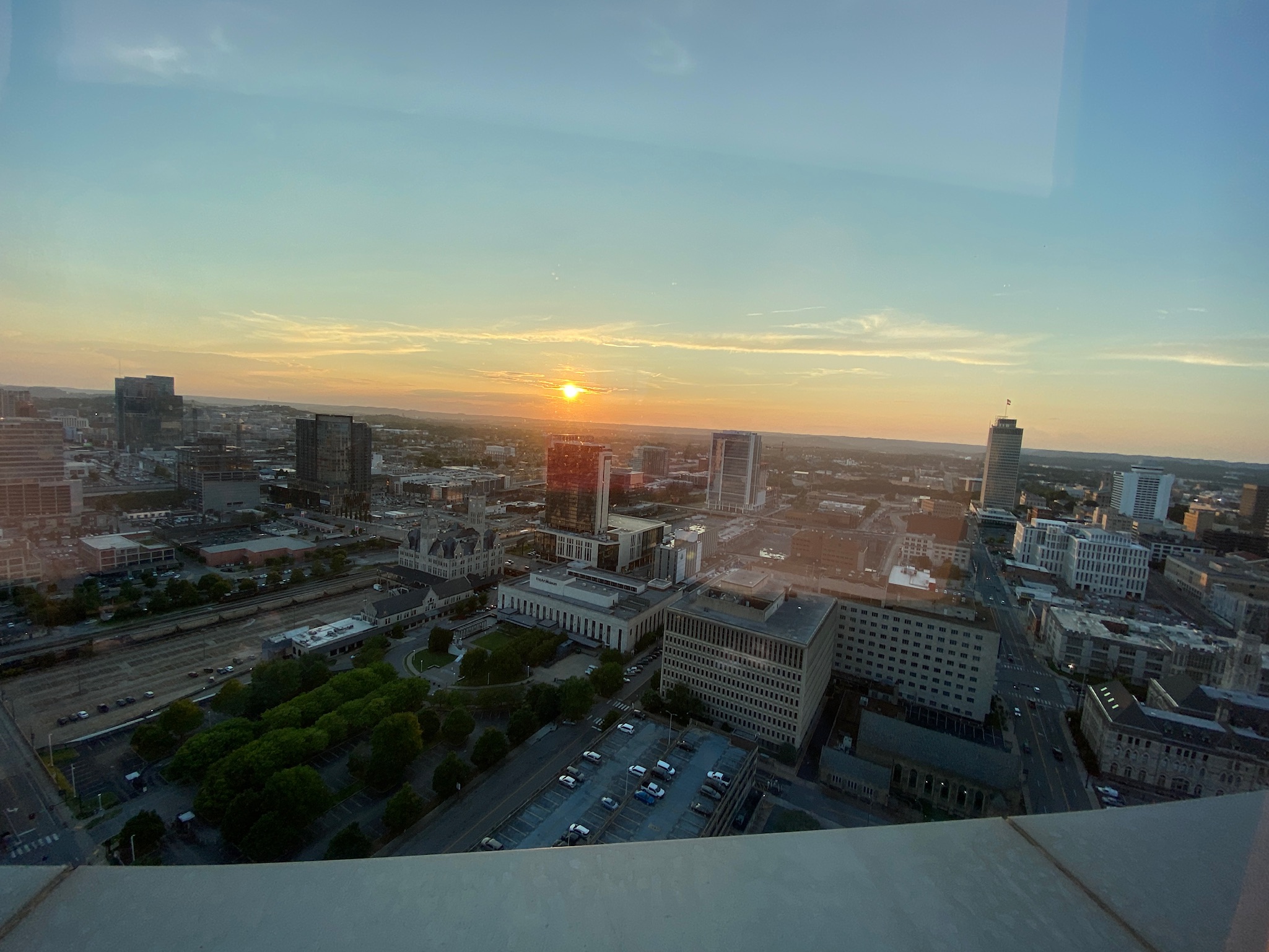 A city skyline at sunset with buildings, roads, and parked cars visible under a clear sky.