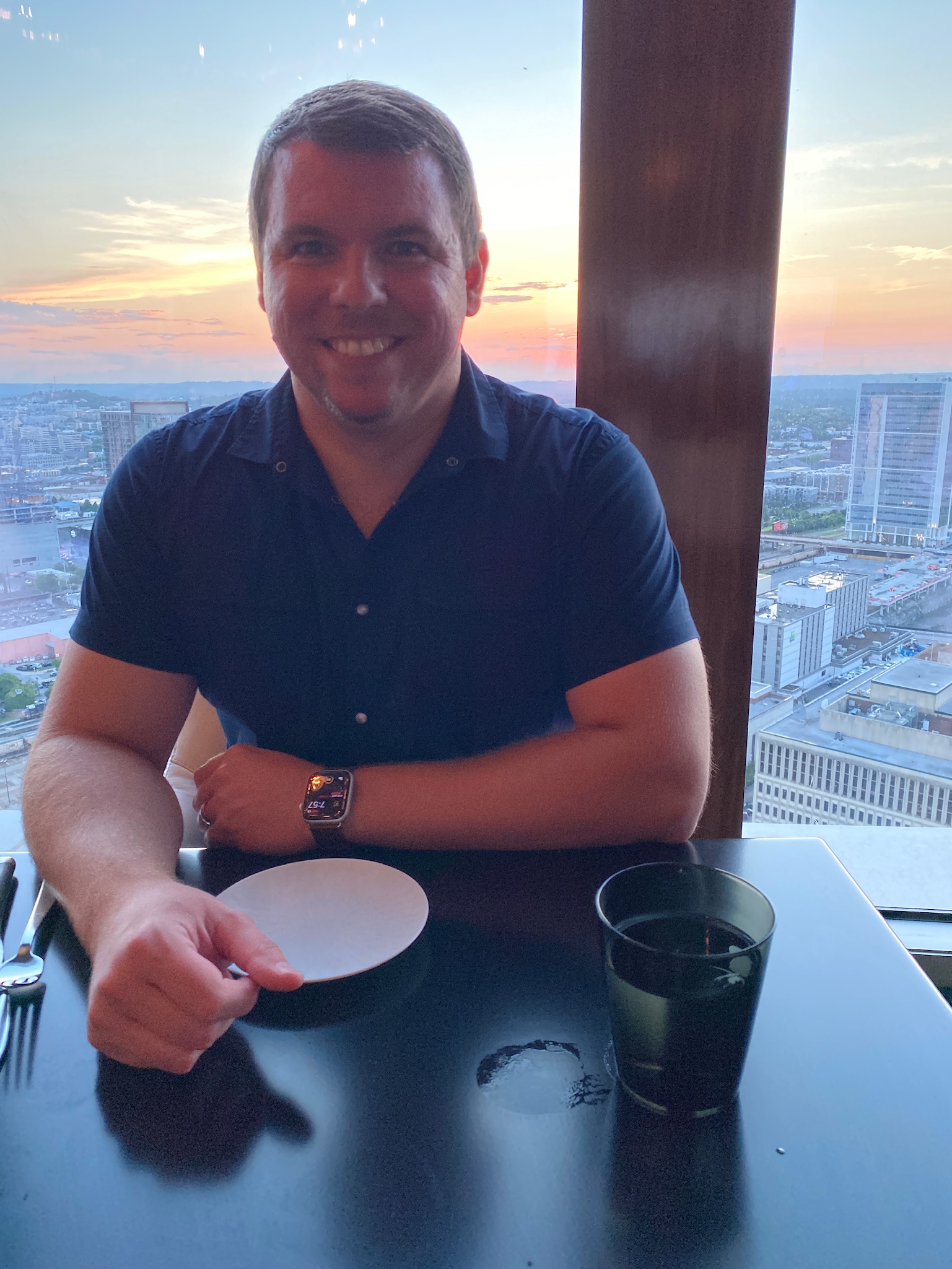 A man sits at a table in a restaurant with a city view at sunset, smiling, with an empty plate, utensils, and a glass of water in front of him.