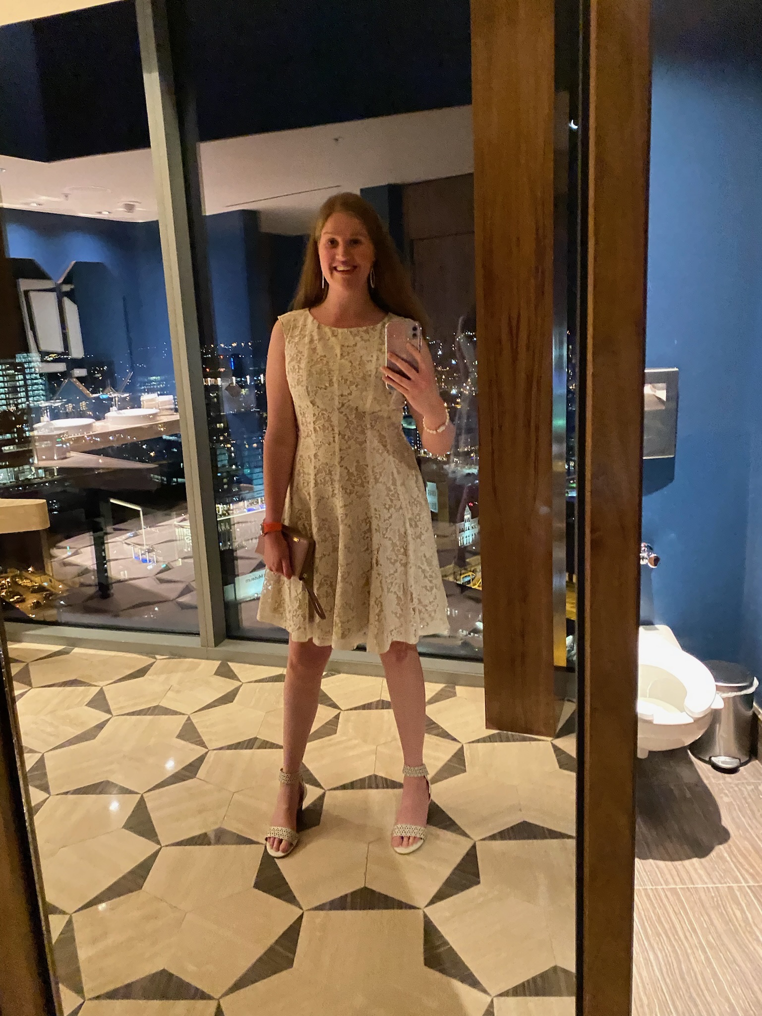 A woman in a white lace dress and heels takes a mirror selfie in a modern, well-lit room with large windows overlooking a city at night.