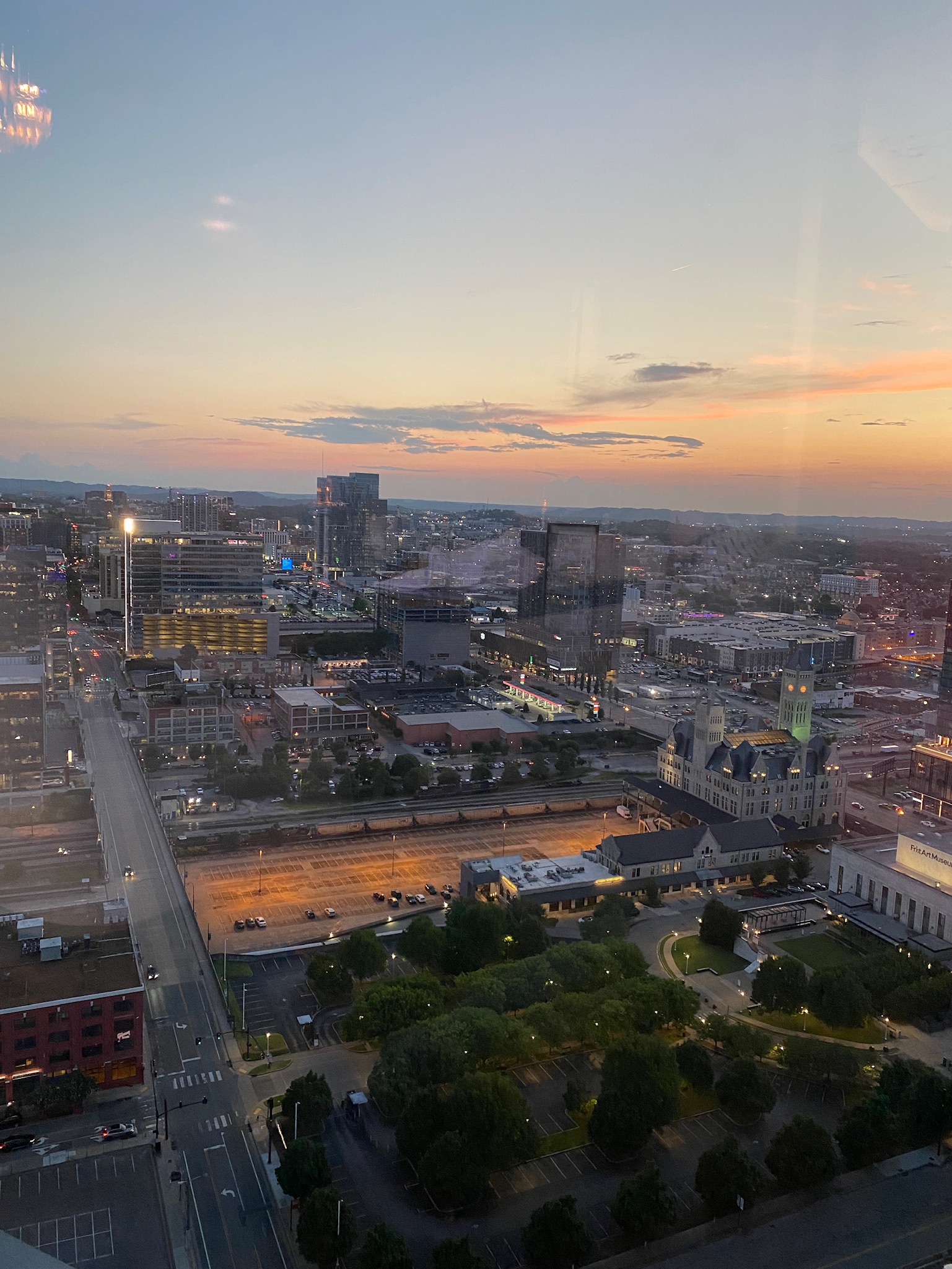 A cityscape at dusk, viewed from a high vantage point, shows buildings, streets, cars, and lights with a colorful sunset in the sky.