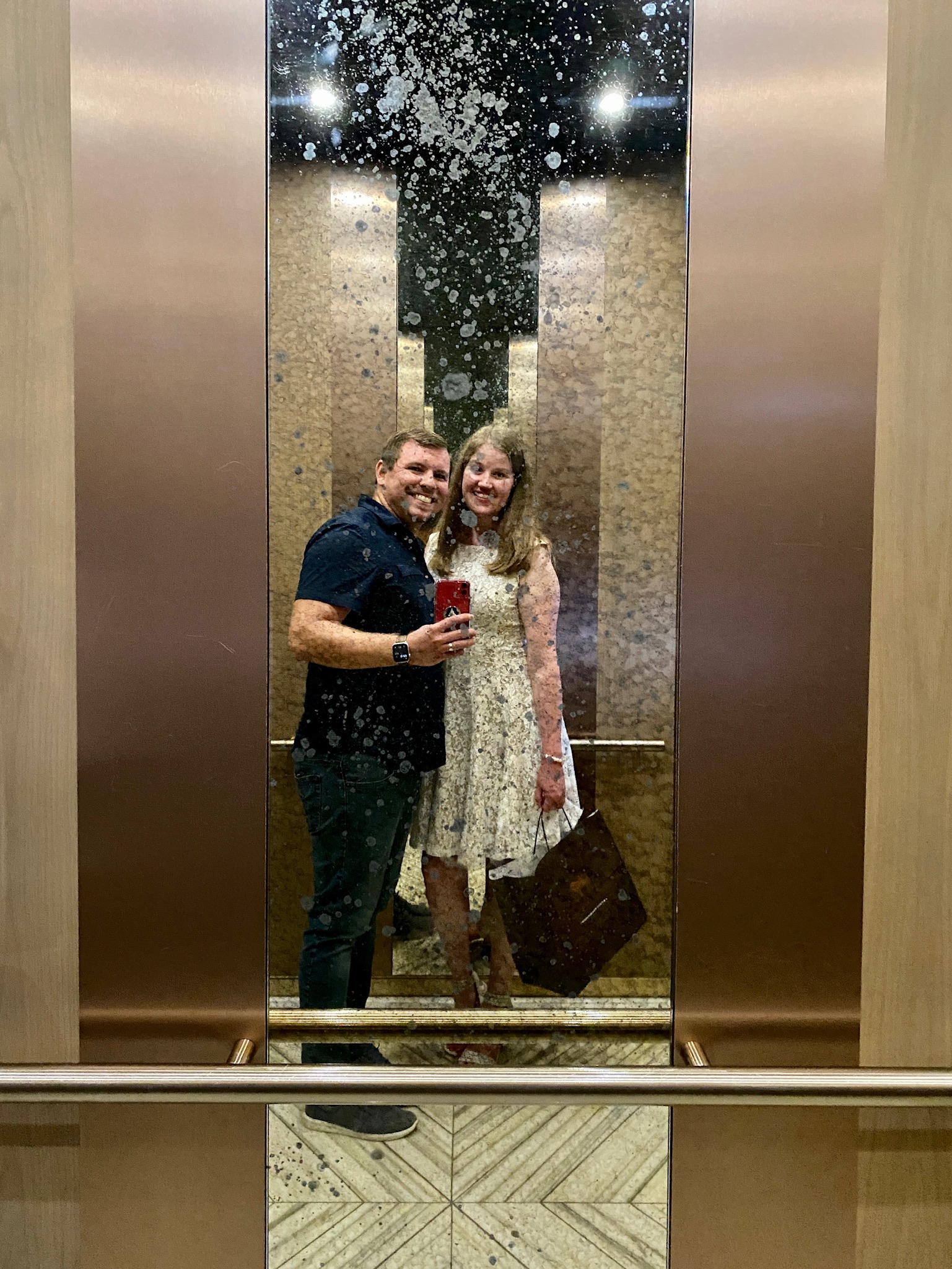 A man and woman take a selfie together in a mirrored elevator. The mirror has visible spots and marks, and the woman holds a large brown bag.