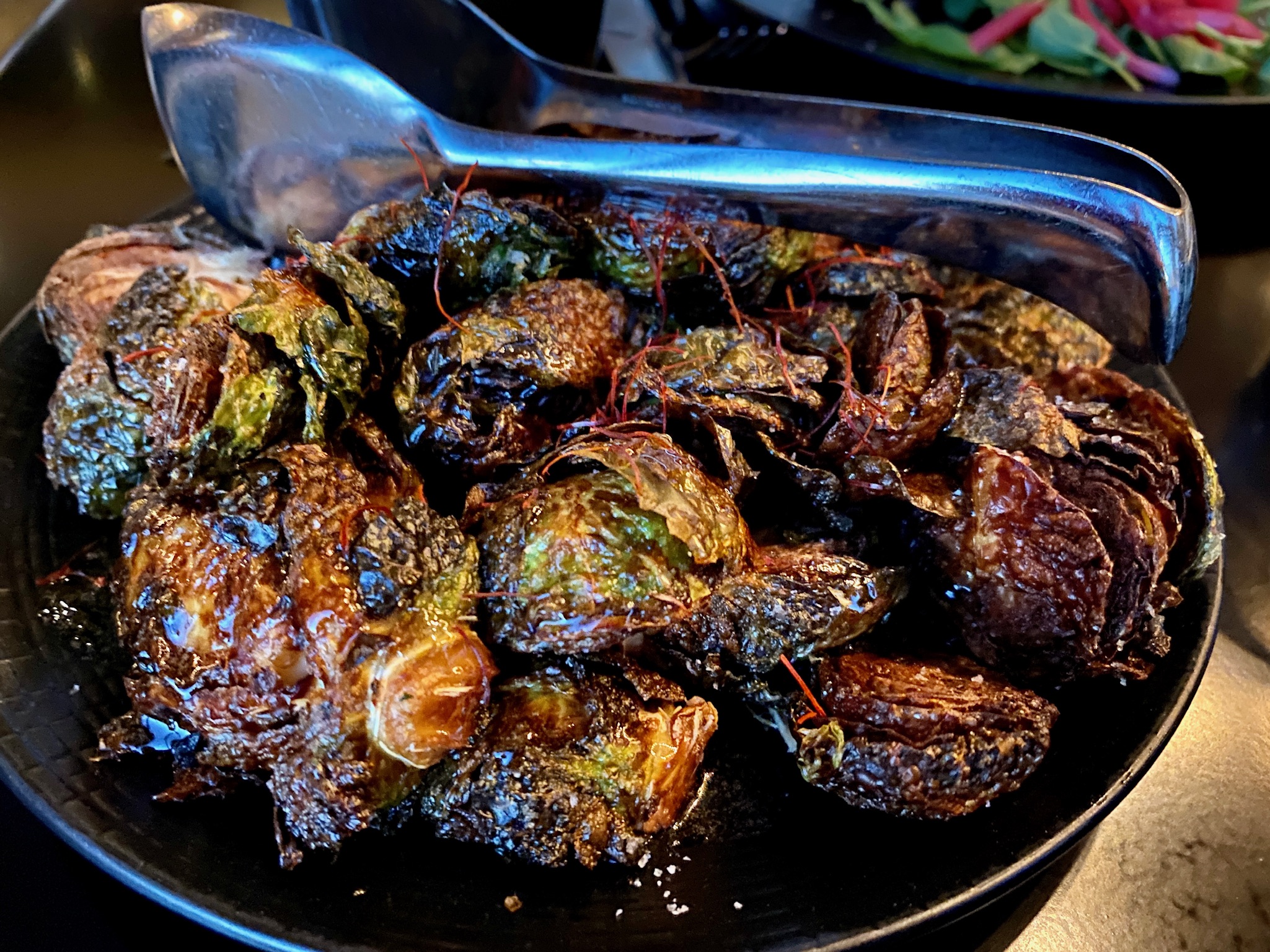 A black plate of roasted Brussels sprouts with crispy, browned edges, served with metal tongs.