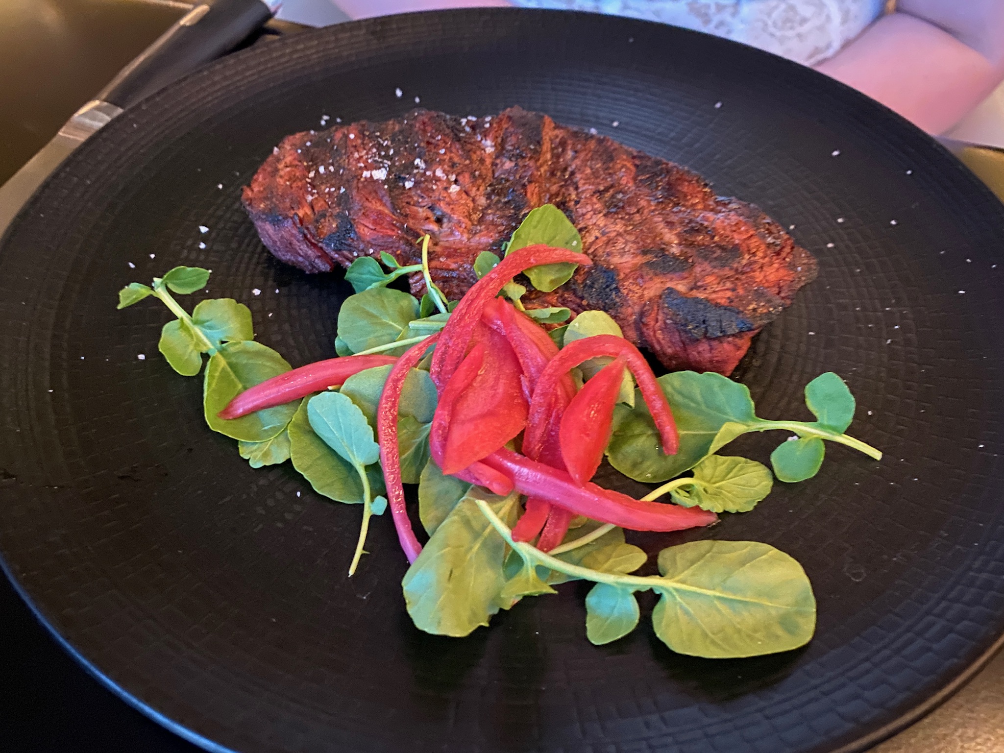 A grilled steak garnished with coarse salt, served with leafy greens and pickled red onions on a black plate.