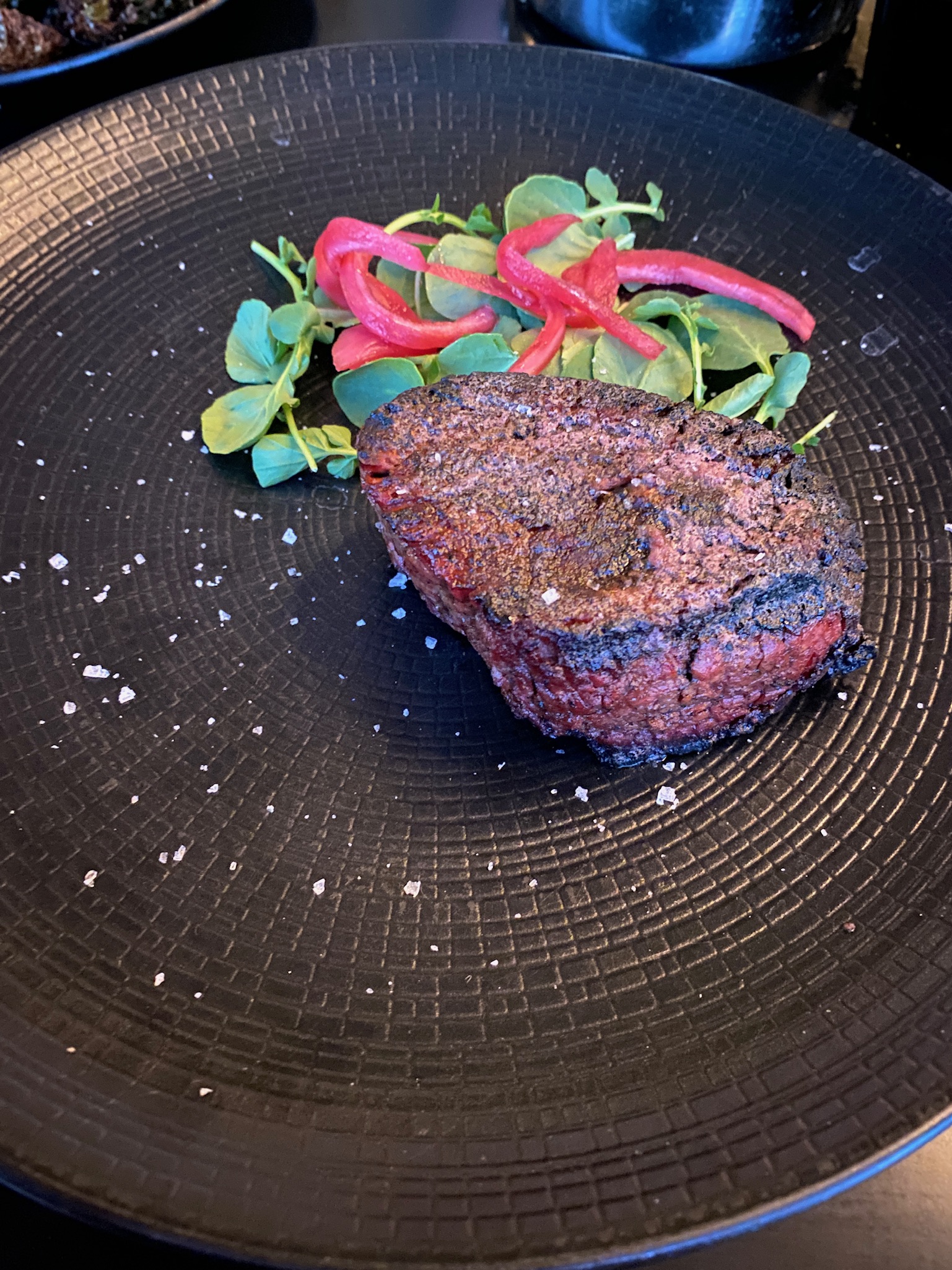 A seared filet mignon steak served on a black plate with microgreens and sliced pickled red onions, garnished with coarse salt.