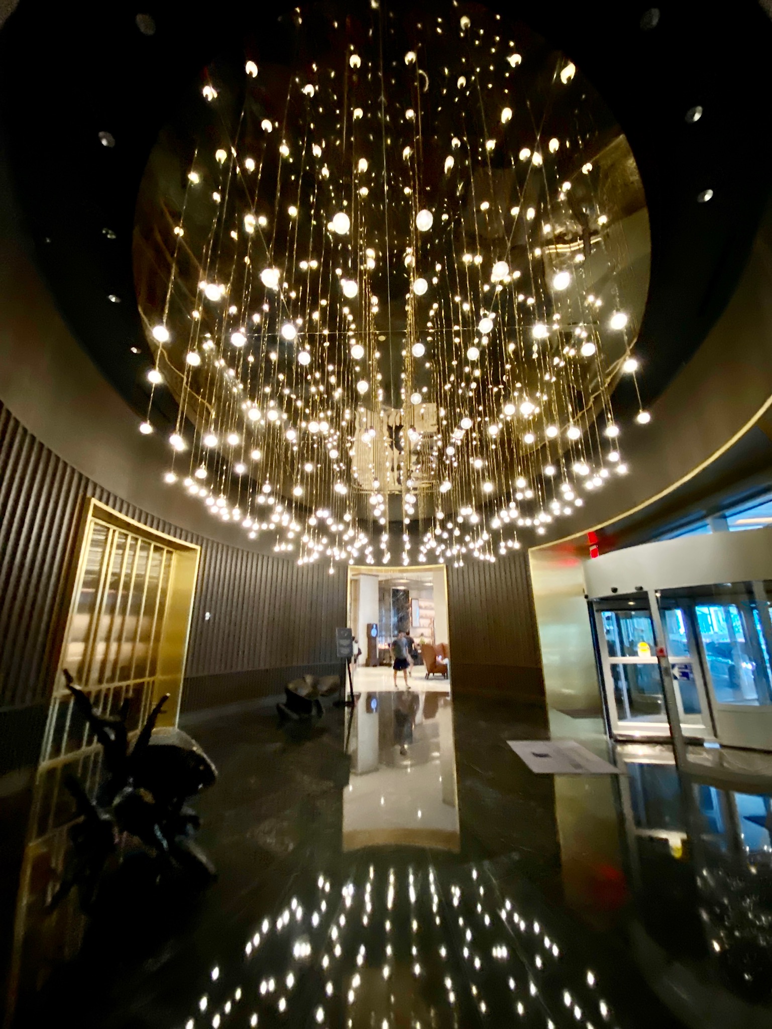 A modern building lobby with a large circular chandelier of hanging lights, shiny black floor reflecting the lights, gold elevator doors, and a glass revolving door entrance.