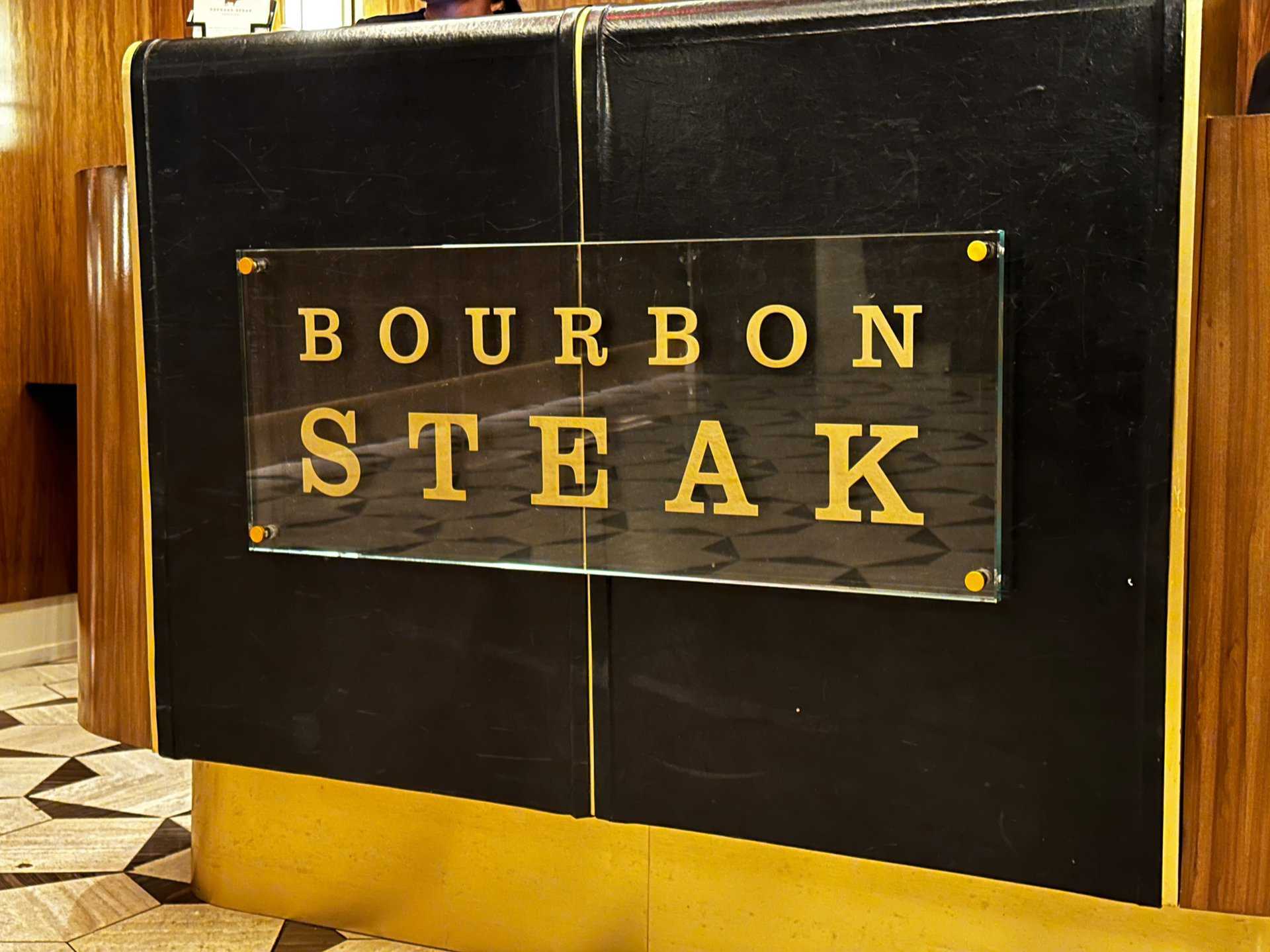 A "Bourbon Steak" sign, Nashville-inspired, graces a dark panel with gold trim, mounted on wood. The geometric floor pattern in the background adds a touch of Southern elegance.