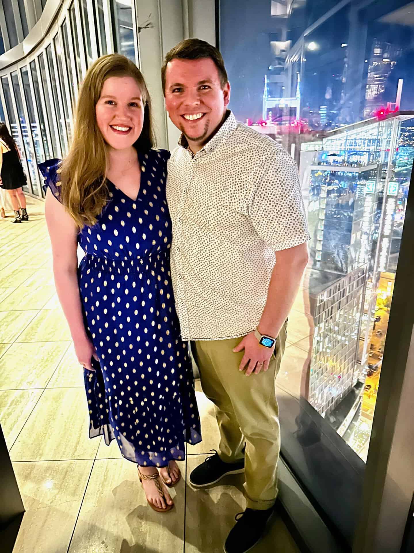 A woman in a blue polka dot dress and a man in a light shirt and khaki pants stand smiling by a large window, overlooking the dazzling Nashville cityscape at night.