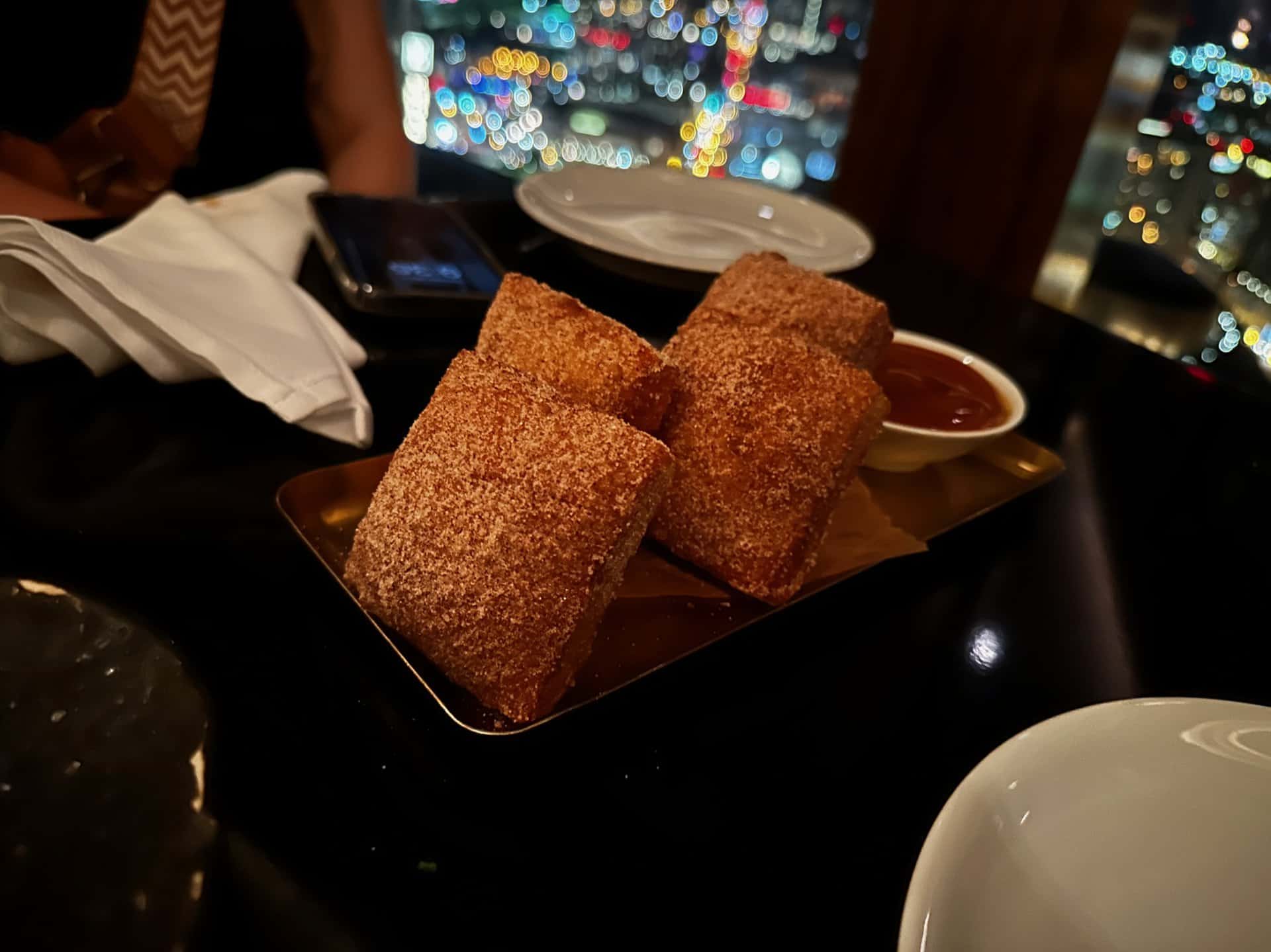A plate with four sugar-coated pastries and a small bowl of dipping sauce sit invitingly on the table, while the vibrant city lights of Nashville twinkle in the background.