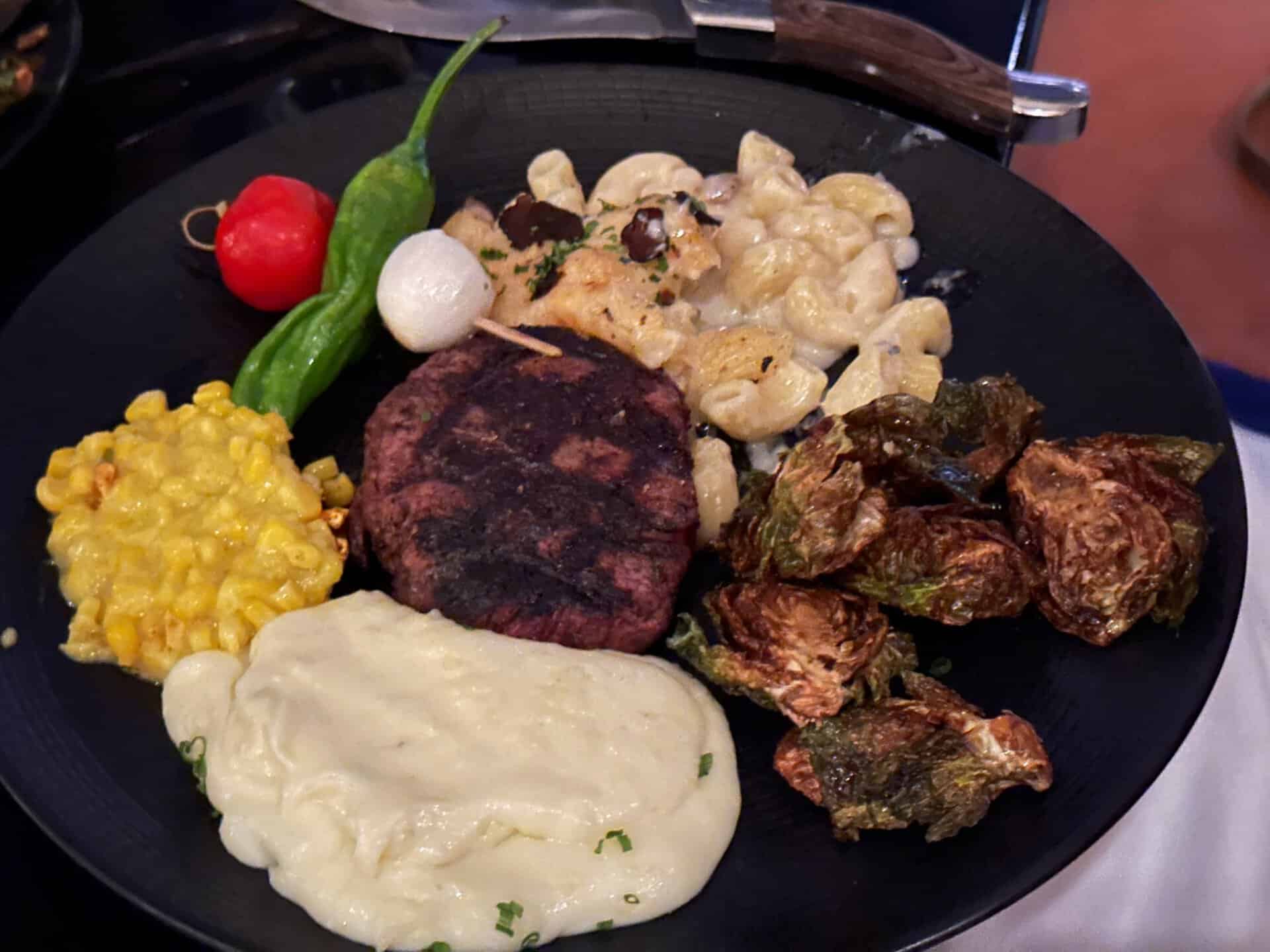 A Nashville-inspired plate with grilled steak, mac and cheese, brussels sprouts, mashed potatoes, corn, a green chili, cherry tomato, and a pearl onion.