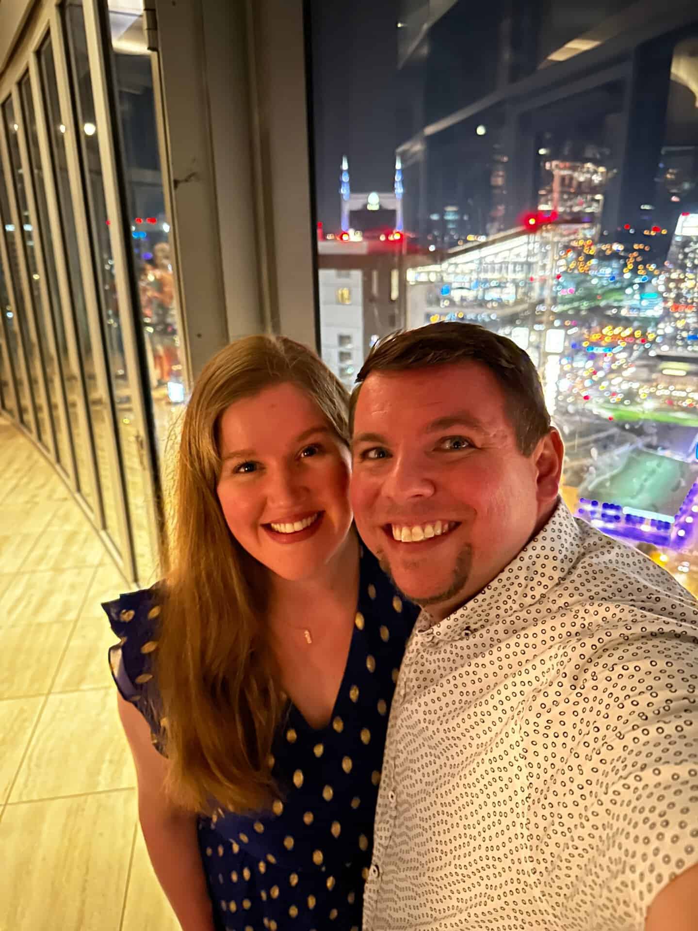 A smiling couple takes a selfie in front of a large window overlooking the vibrant Nashville cityscape, its bright lights illuminating the night.