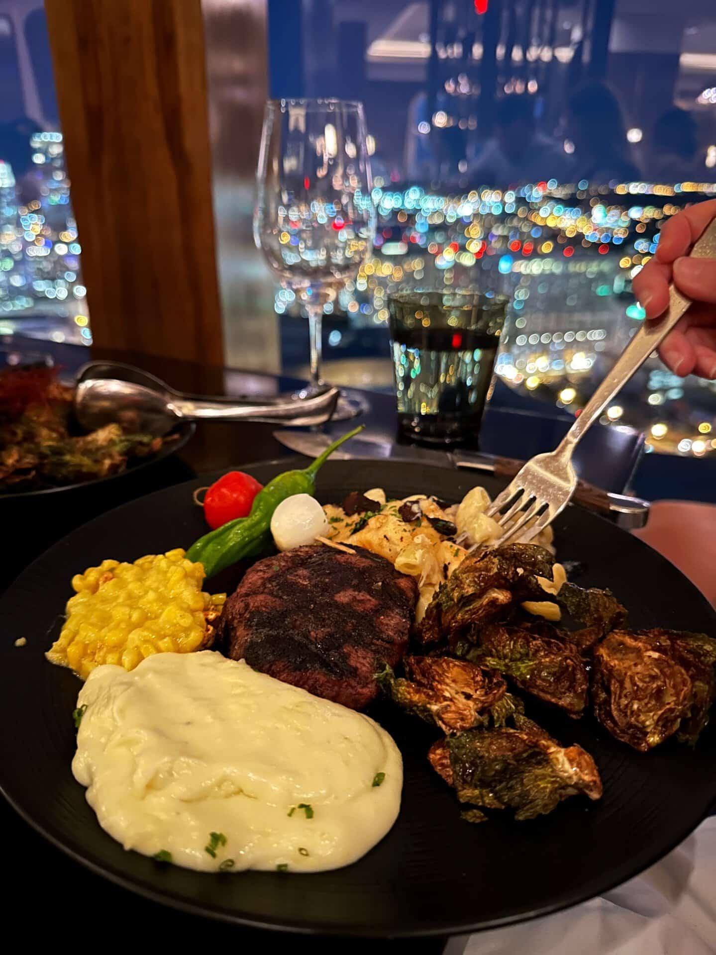 A plate of steak, mashed potatoes, corn, Brussels sprouts, and vegetables sits against a stunning Nashville cityscape view through the window. A fork is poised over the plate.
