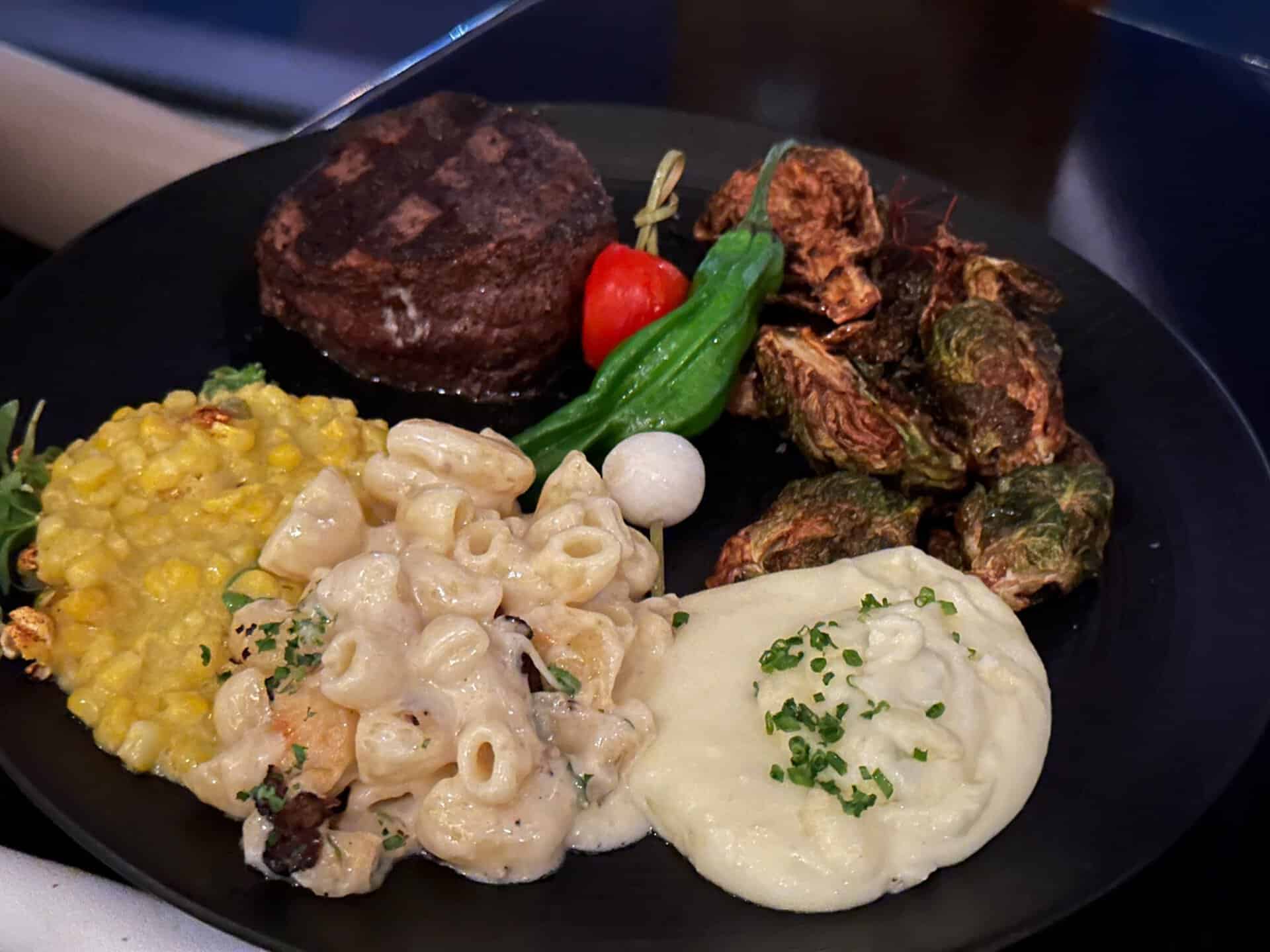 A plate featuring Nashville-inspired steak, grilled tomato, green peppers, crispy Brussels sprouts, macaroni and cheese, creamed corn, and mashed potatoes garnished with chives.