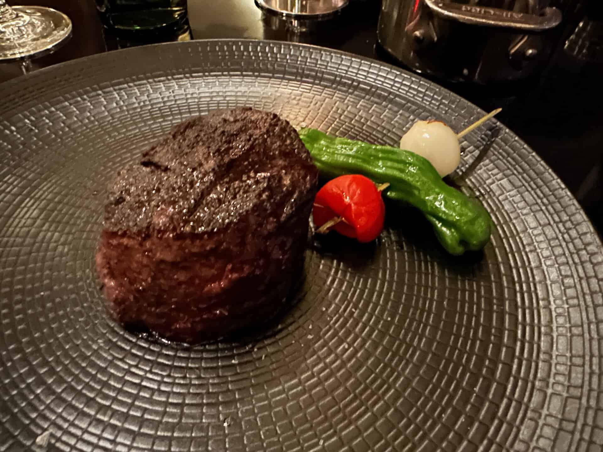 A Nashville-inspired cooked filet steak is elegantly served on a textured black plate, complemented by a skewer holding a small onion, green pepper, and cherry tomato.