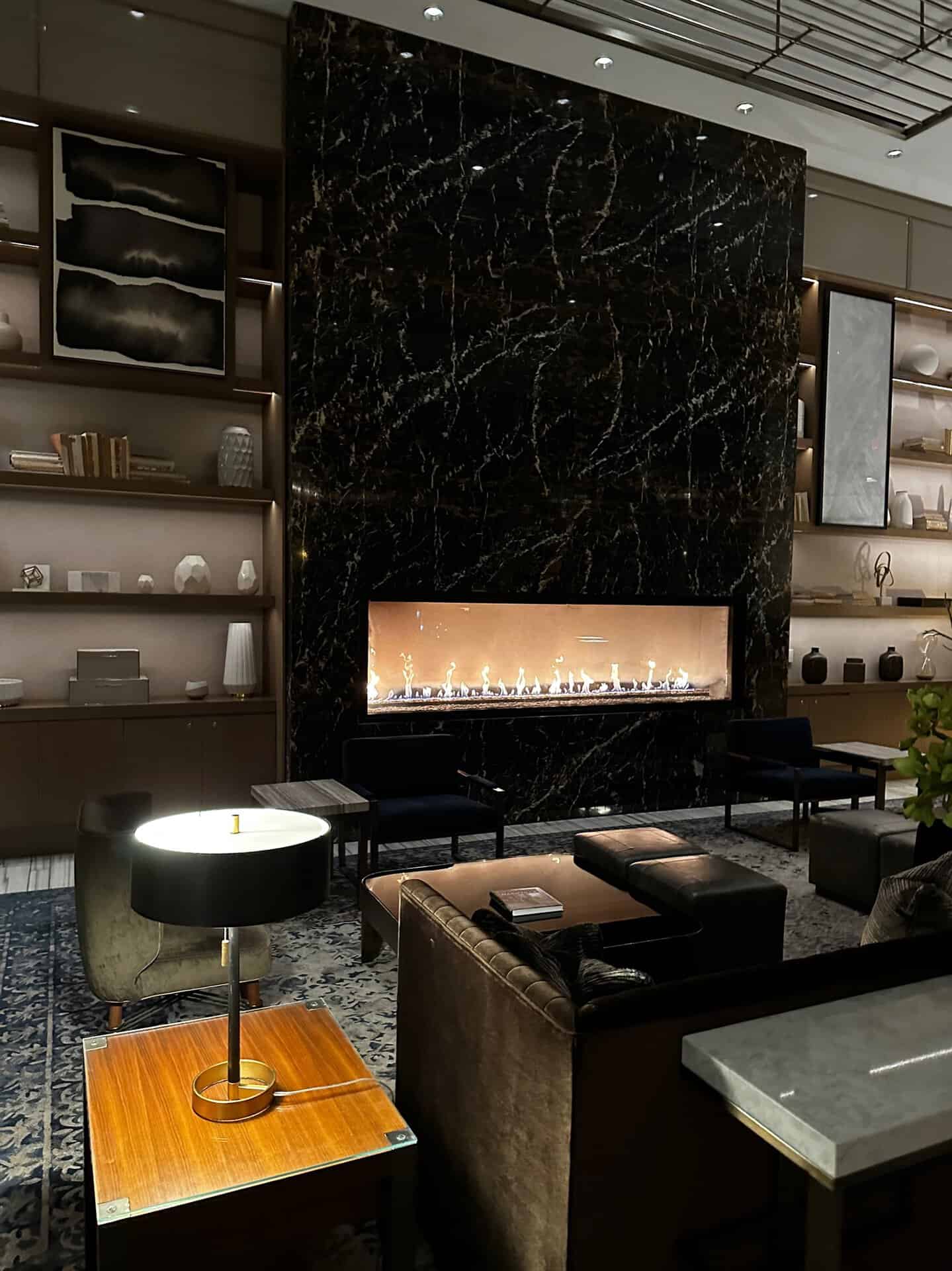 Nashville-inspired modern lounge area featuring a long marble fireplace, stylish chairs, and a wooden table with a lamp. Dim lighting highlights the contemporary design, while shelves adorned with decor add to the room's inviting atmosphere.