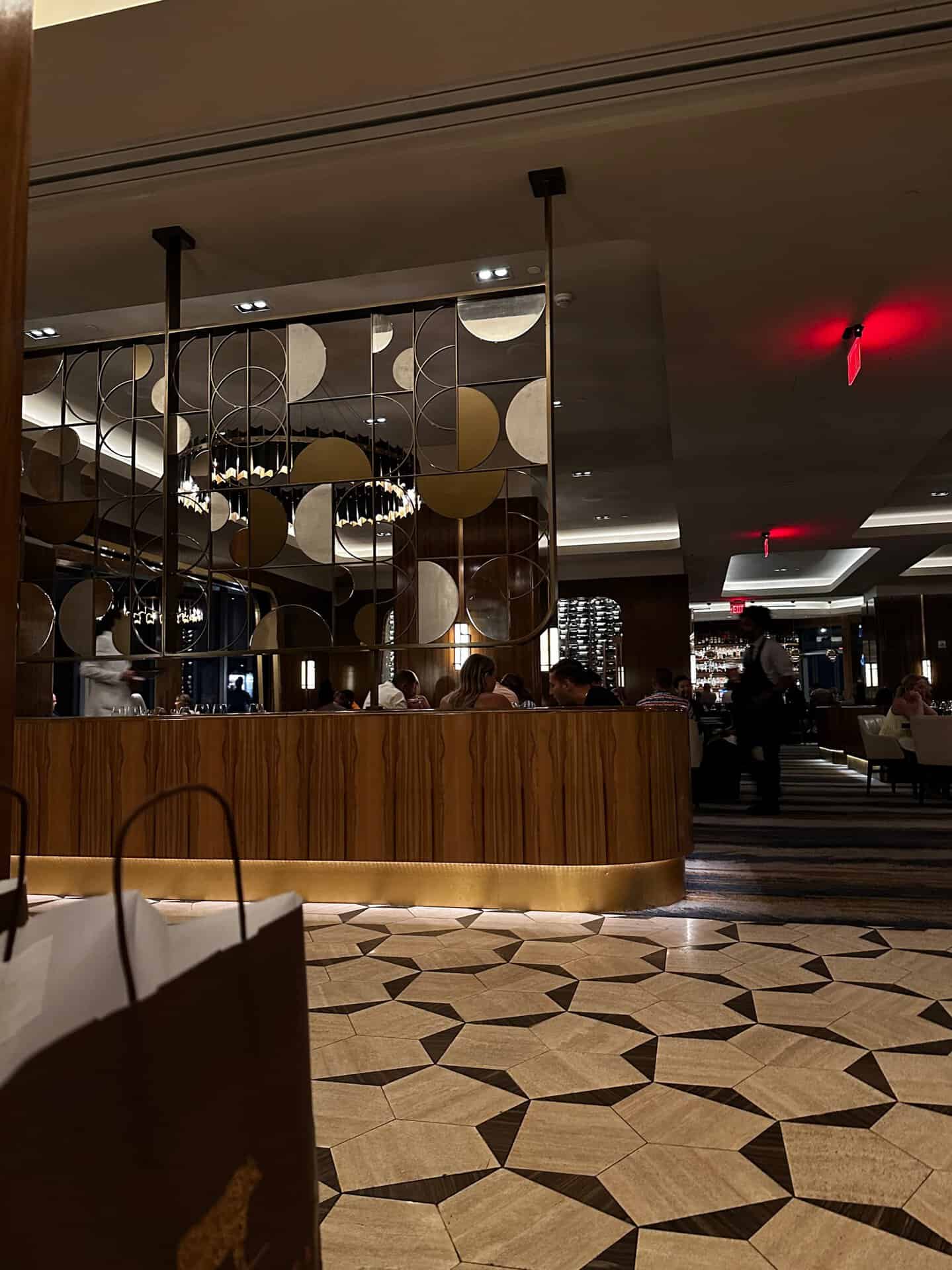 The dimly lit Nashville restaurant features a geometric patterned floor, modern circular wall decor, and tables of seated patrons. A waiter moves in the background while a paper bag sits prominently in the foreground, adding to the scene's charm.