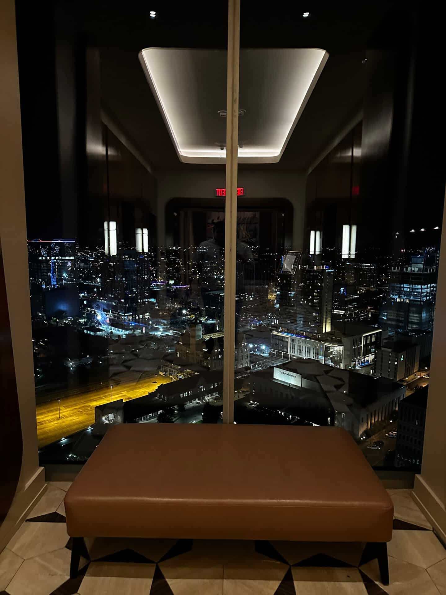 Nashville's night cityscape unfurls beyond the large window, where buildings and streetlights twinkle in harmony. A brown bench sits gracefully in the dimly lit room, offering a perfect spot to ponder the urban beauty.