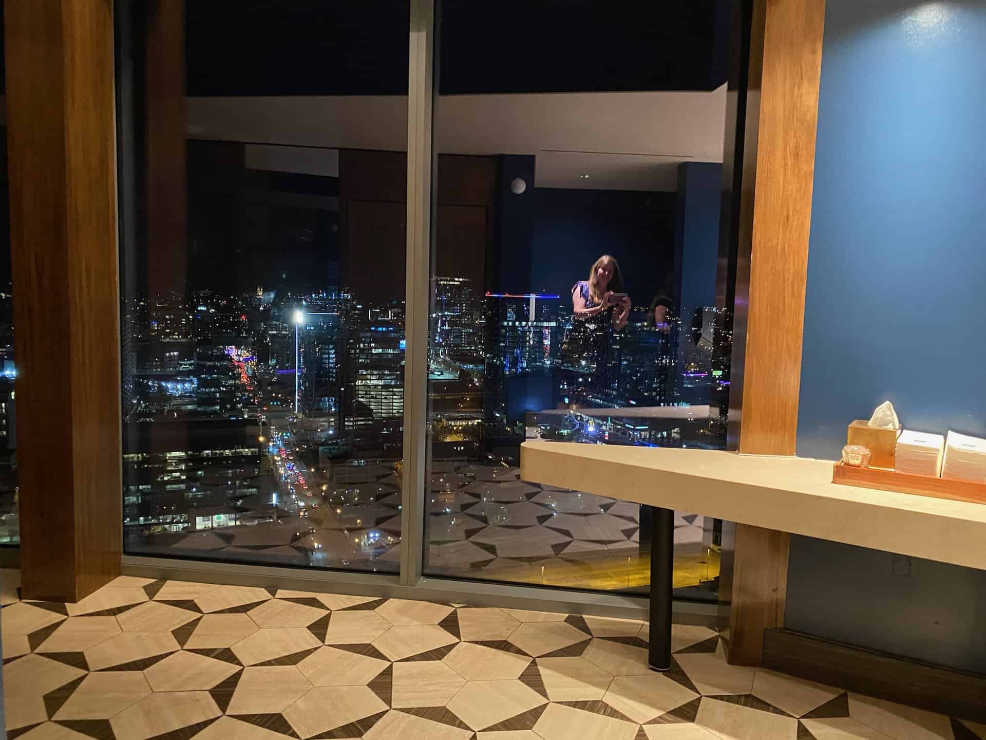 City skyline view of Nashville from a high-rise apartment with floor-to-ceiling windows. The indoor scene features a geometric-patterned floor and a modern table adorned with a tissue box.