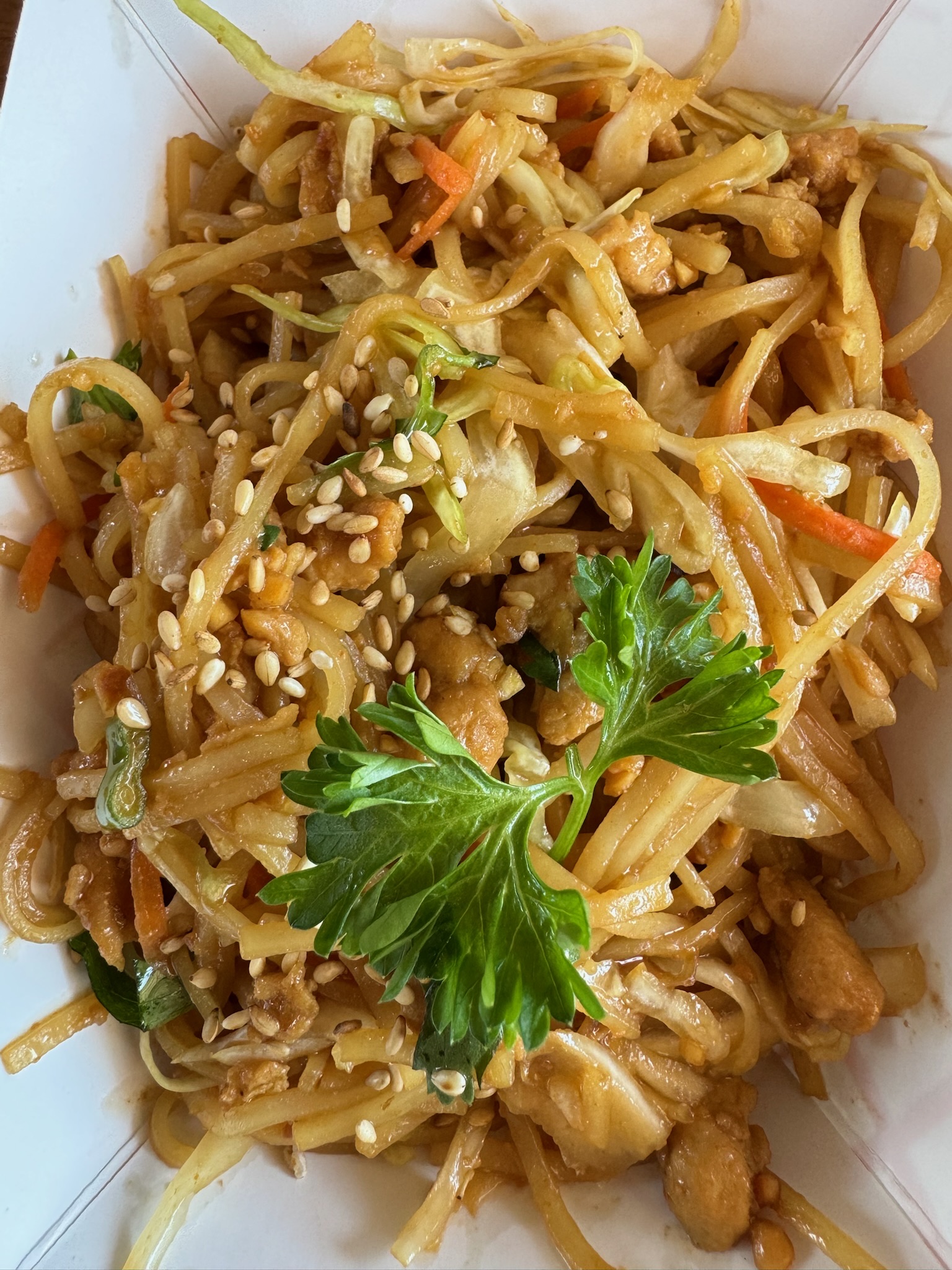 A close-up of stir-fried noodles with vegetables, diced chicken, sesame seeds, and a sprig of parsley served in a white takeout container.