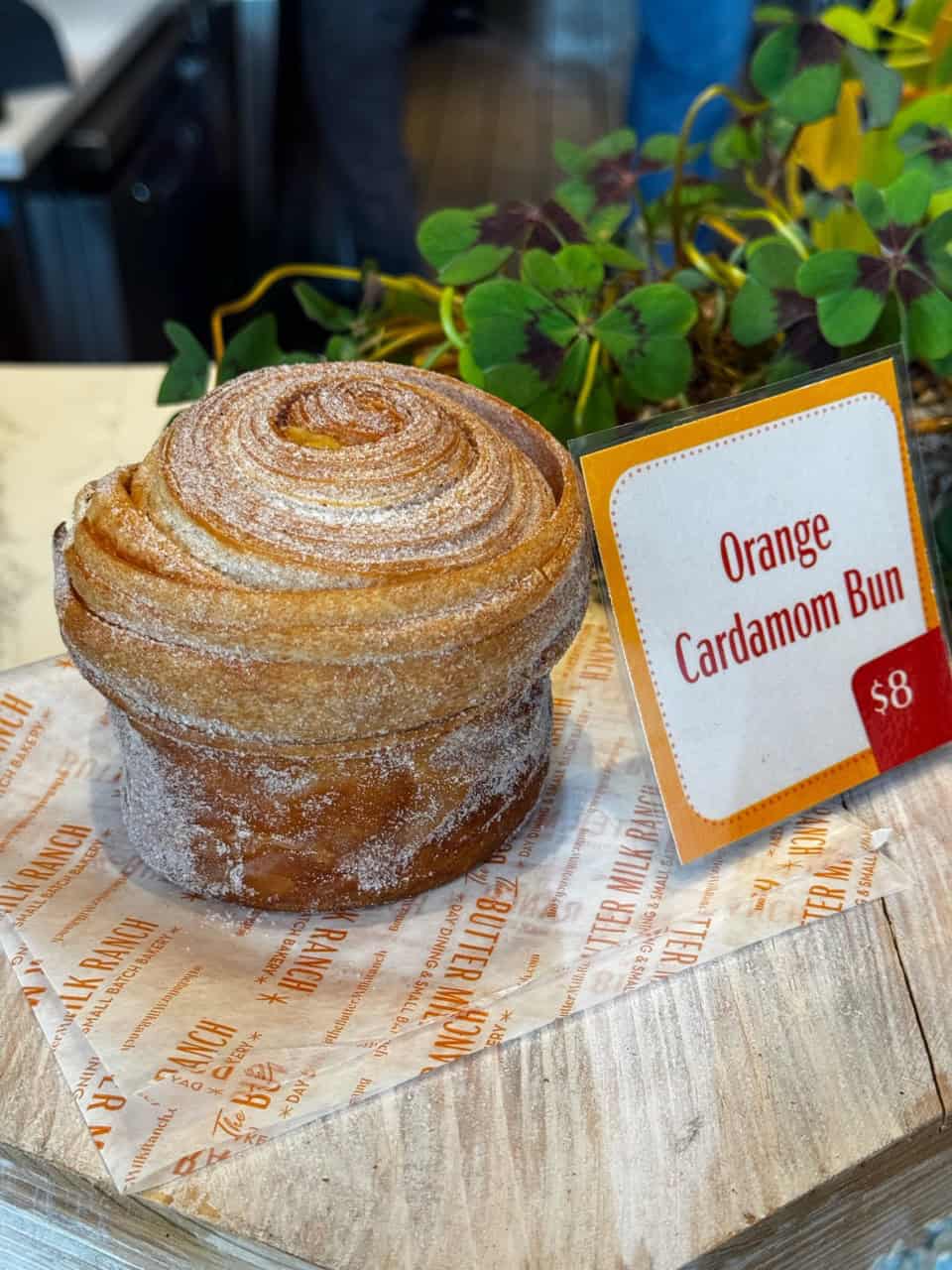 A swirled Orange Cardamom Bun rests on a wooden block, priced at $8, set against the charming backdrop of Nashville's artisan cafes, with a small plant peeking into the scene.