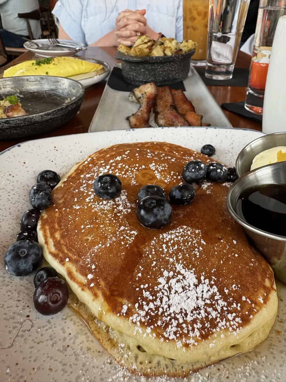 A plate of pancakes topped with Nashville blueberries and powdered sugar, syrup on the side. In the background, bacon, potatoes, and an omelette evoke a Southern breakfast feast.
