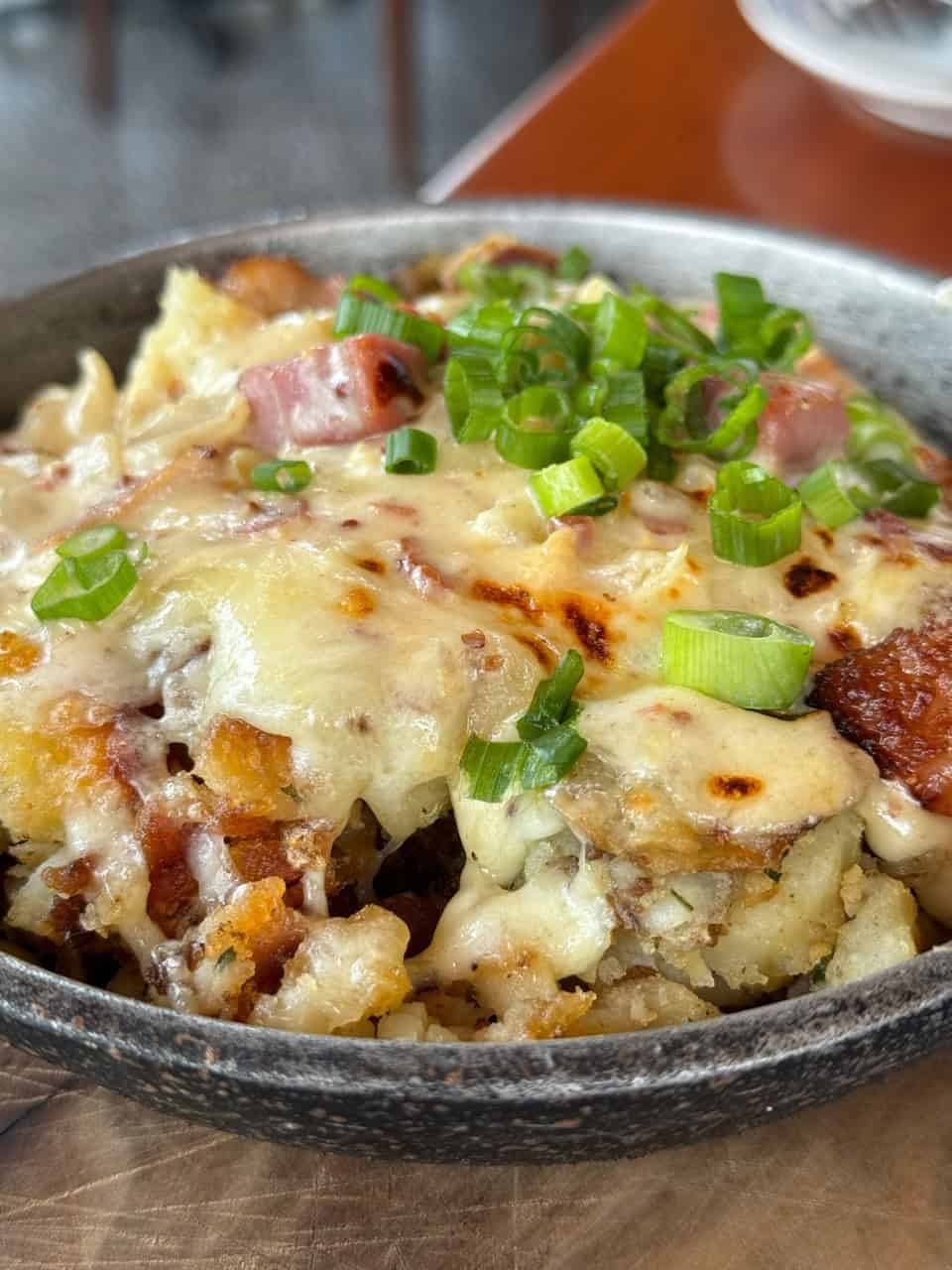 A skillet dish of cheesy potatoes, Nashville-style, topped with diced ham and chopped green onions.