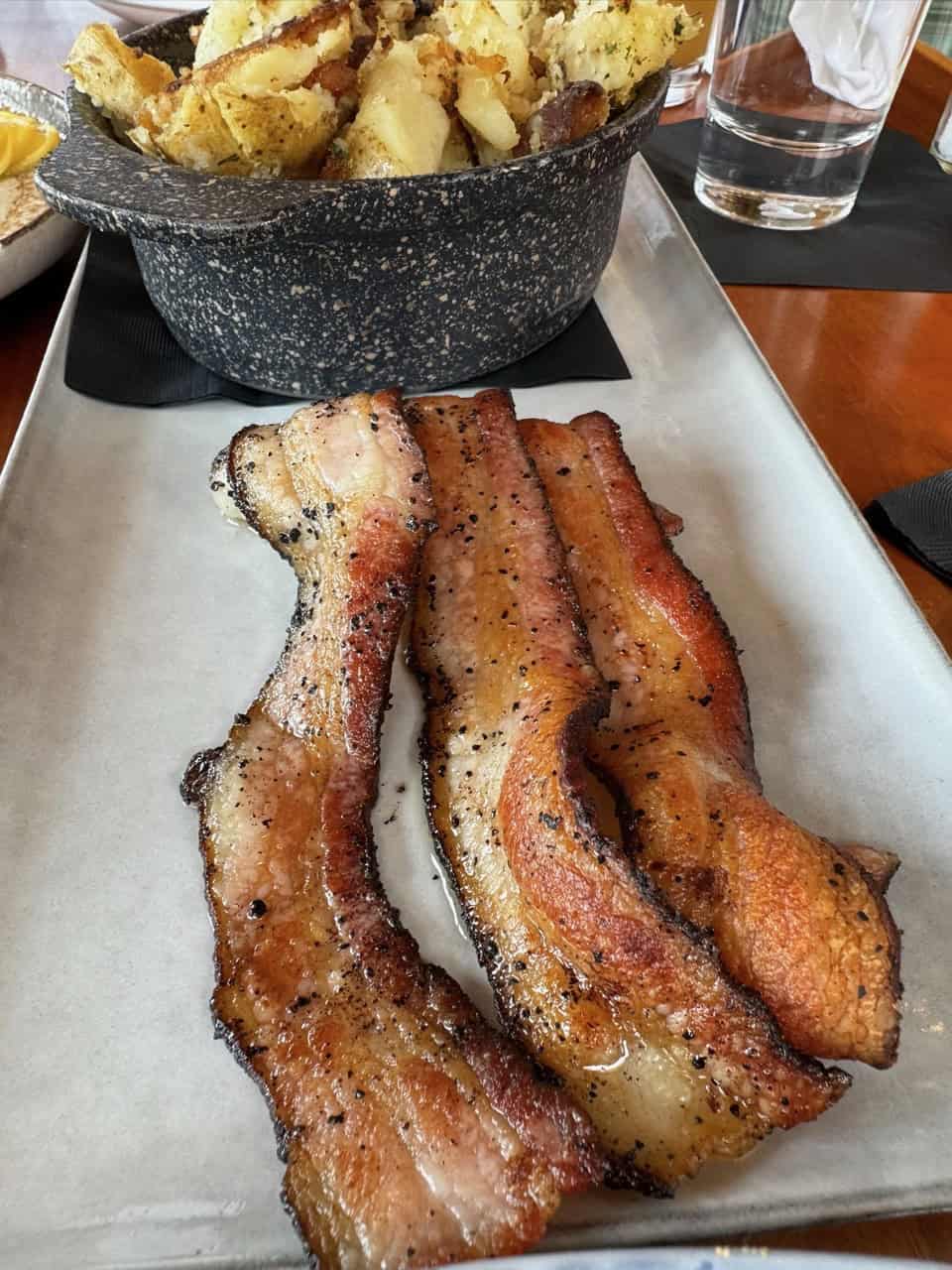 Three slices of cooked bacon rest elegantly on a rectangular plate, with a bowl of roasted potatoes subtly evoking the comforting flavors of Nashville in the background.