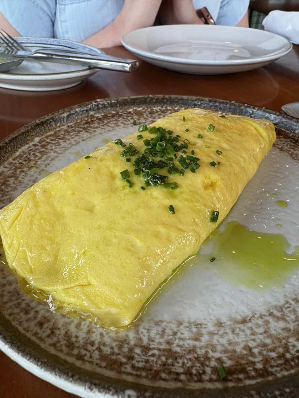 A neatly folded omelette, reminiscent of a Nashville brunch spot, is garnished with chopped chives on a ceramic plate. A person in a blue shirt is partially visible in the background.