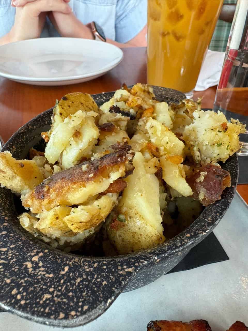 A black bowl filled with roasted potato chunks sits on a Nashville table. A person is seated with hands clasped in anticipation, while a tall glass of iced tea adds a refreshing Southern touch nearby.