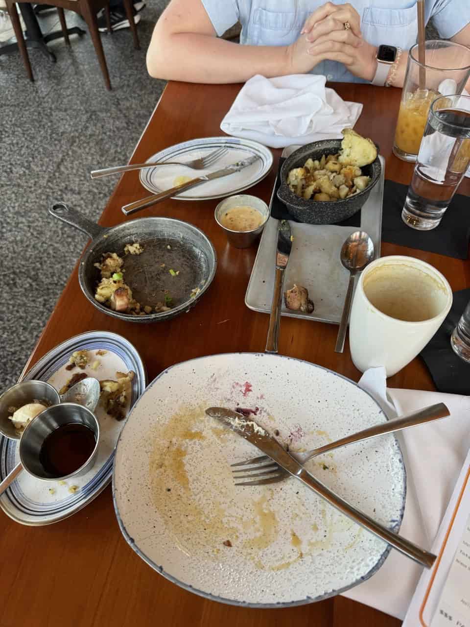 Empty plates and dishes with remnants of Southern cuisine hint at a delightful feast on the table, alongside drinks and utensils. In the backdrop, a person with folded hands enjoys the laid-back vibe characteristic of Nashville.