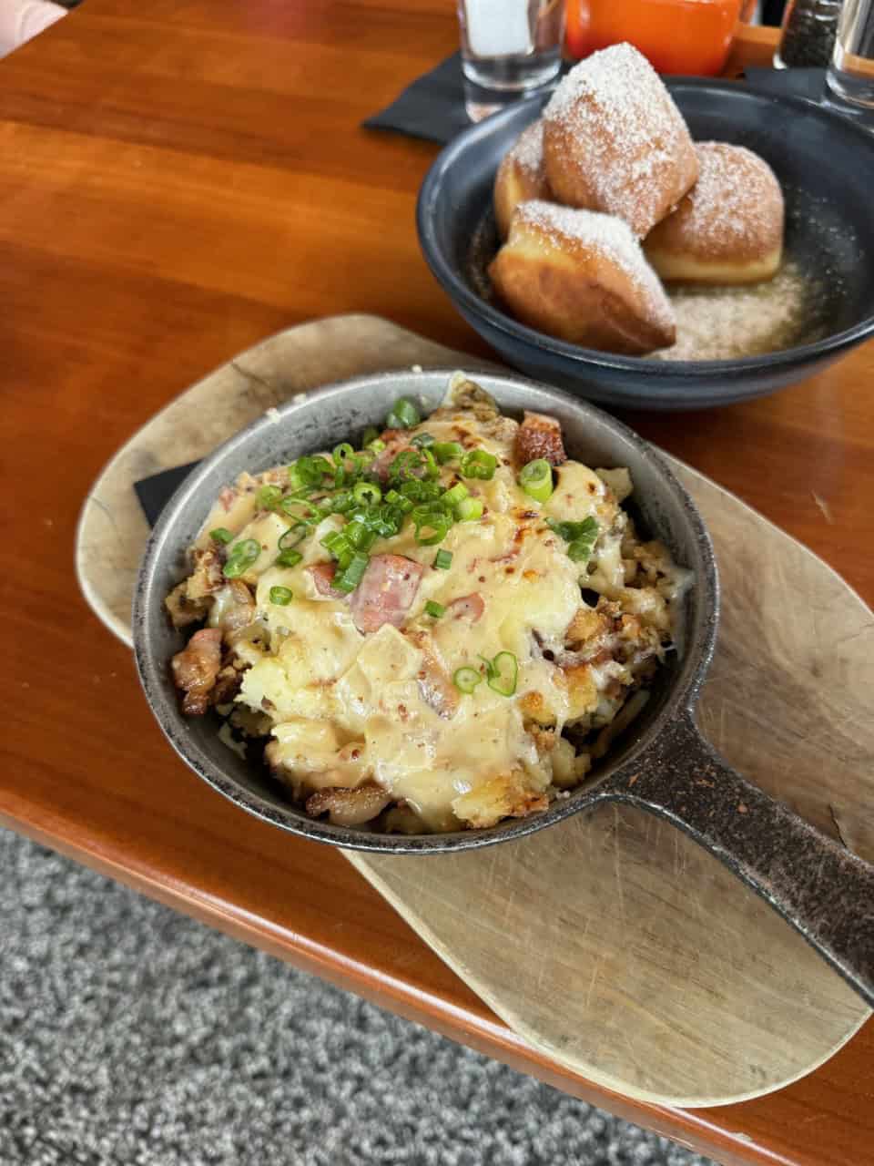 A Nashville-inspired skillet of melted cheese and chopped green onions graces a dish with meat and potatoes, perched on a wooden board. In the background, a black bowl brims with powdered sugar-coated beignets.