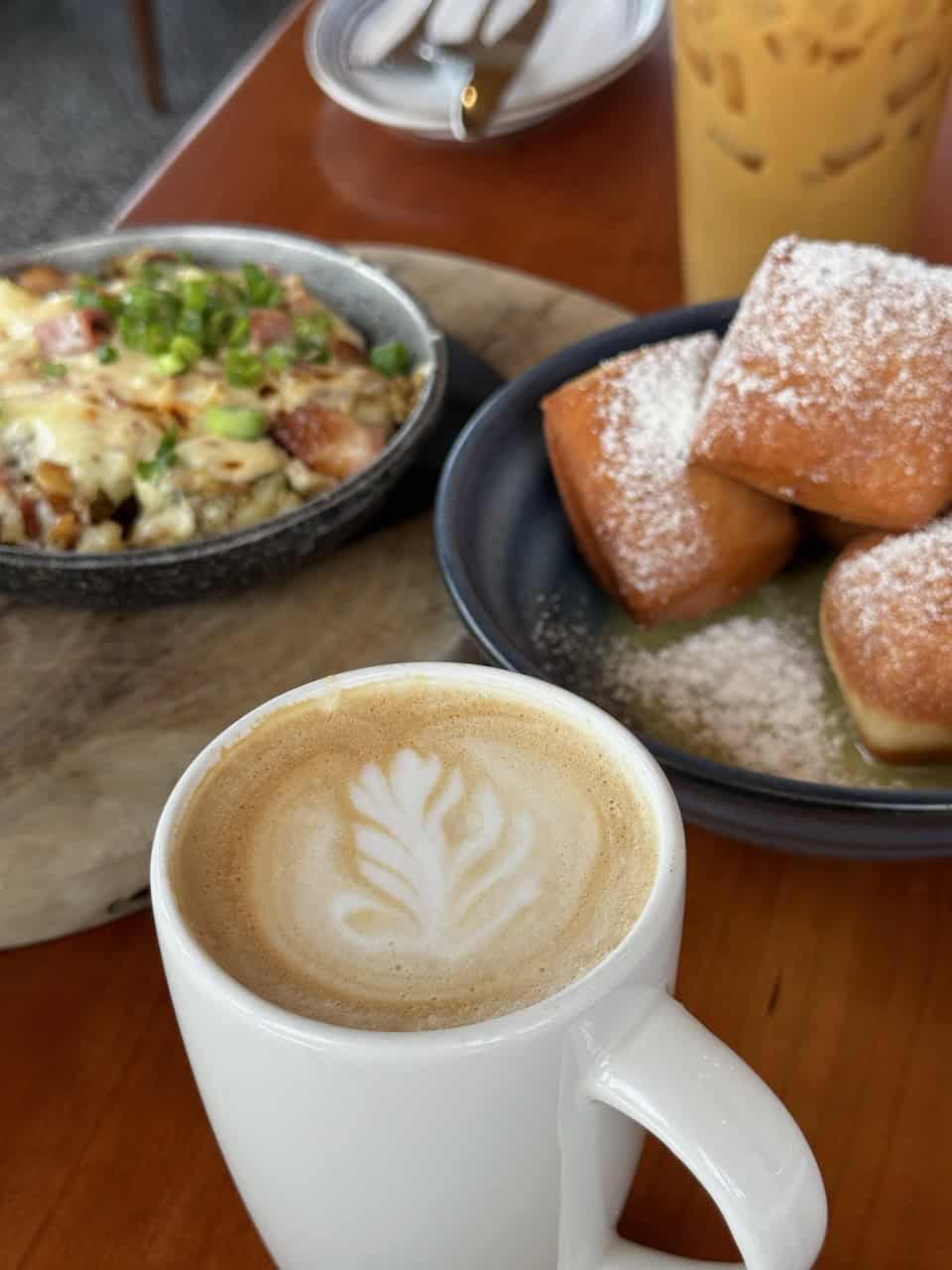 A Nashville-inspired latte with intricate art, a dish topped with melted cheese and scallions, and a plate of powdered sugar-covered pastries all sit invitingly on a rustic wooden table.